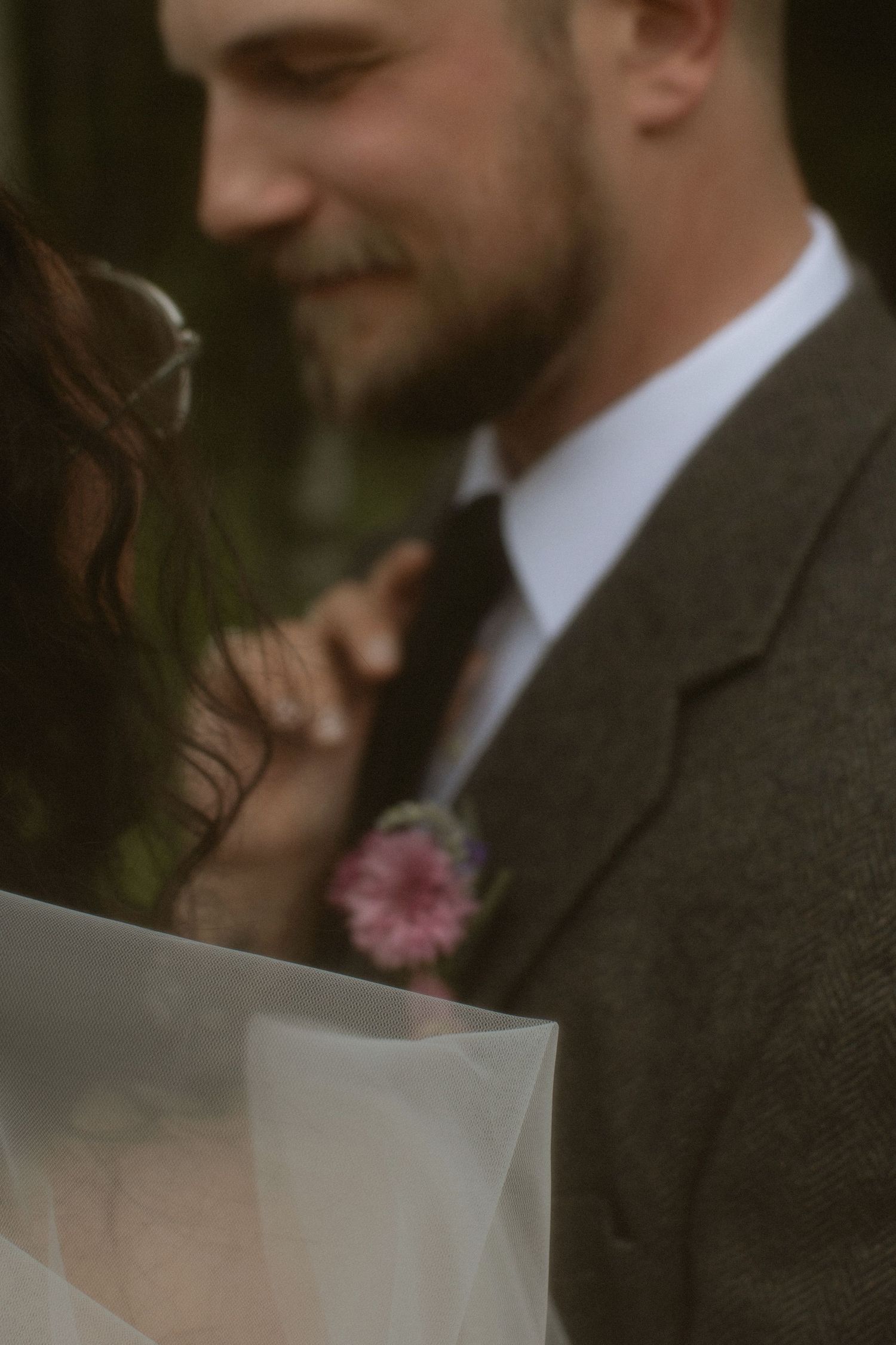 A groom in a brown tweed suit with a pink boutonniere smiles tenderly at his bride during their wedding ceremony.