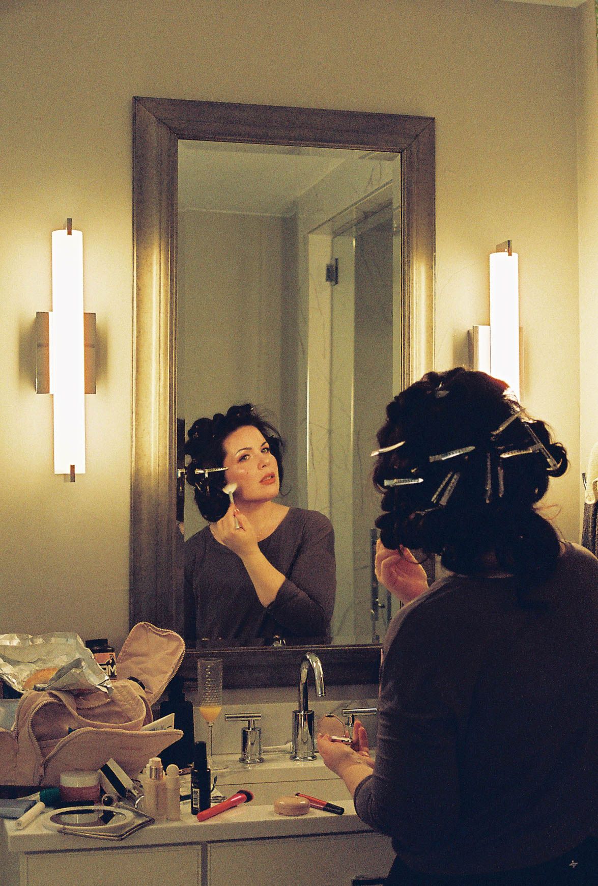 Bride with hair rollers applying makeup at a bathroom mirror before her wedding.