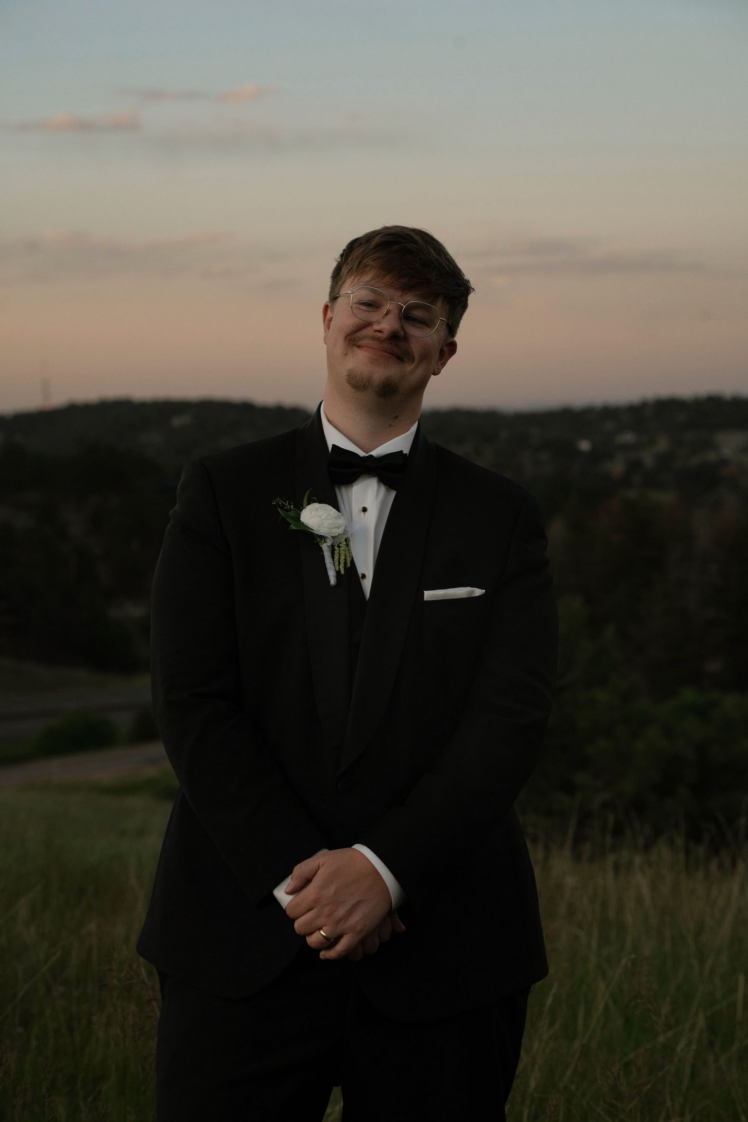 Young man in black tuxedo with white boutonniere and bow tie smiling outdoors at dusk.