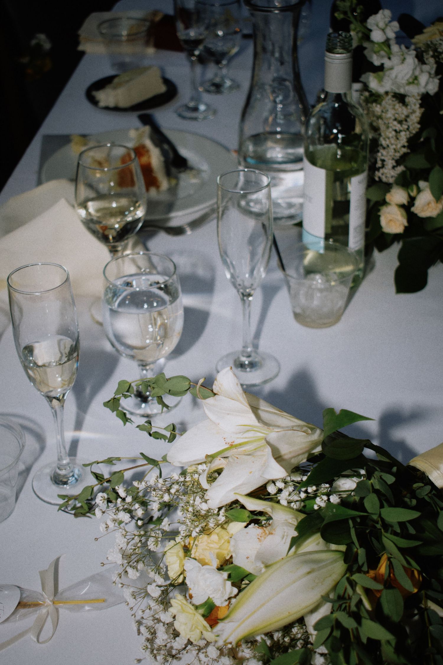 Elegant wedding reception table with white floral bouquet, champagne flutes, wine bottle, and glassware on white linen.