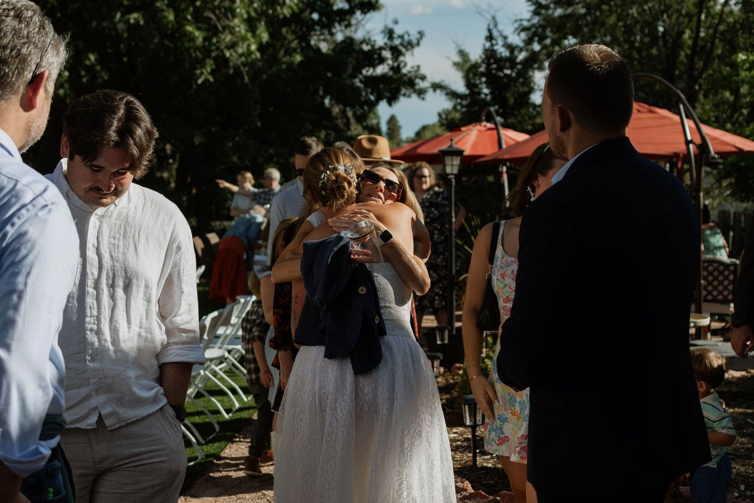 Bride in white dress embracing a guest at an outdoor wedding reception with red umbrellas in the background.