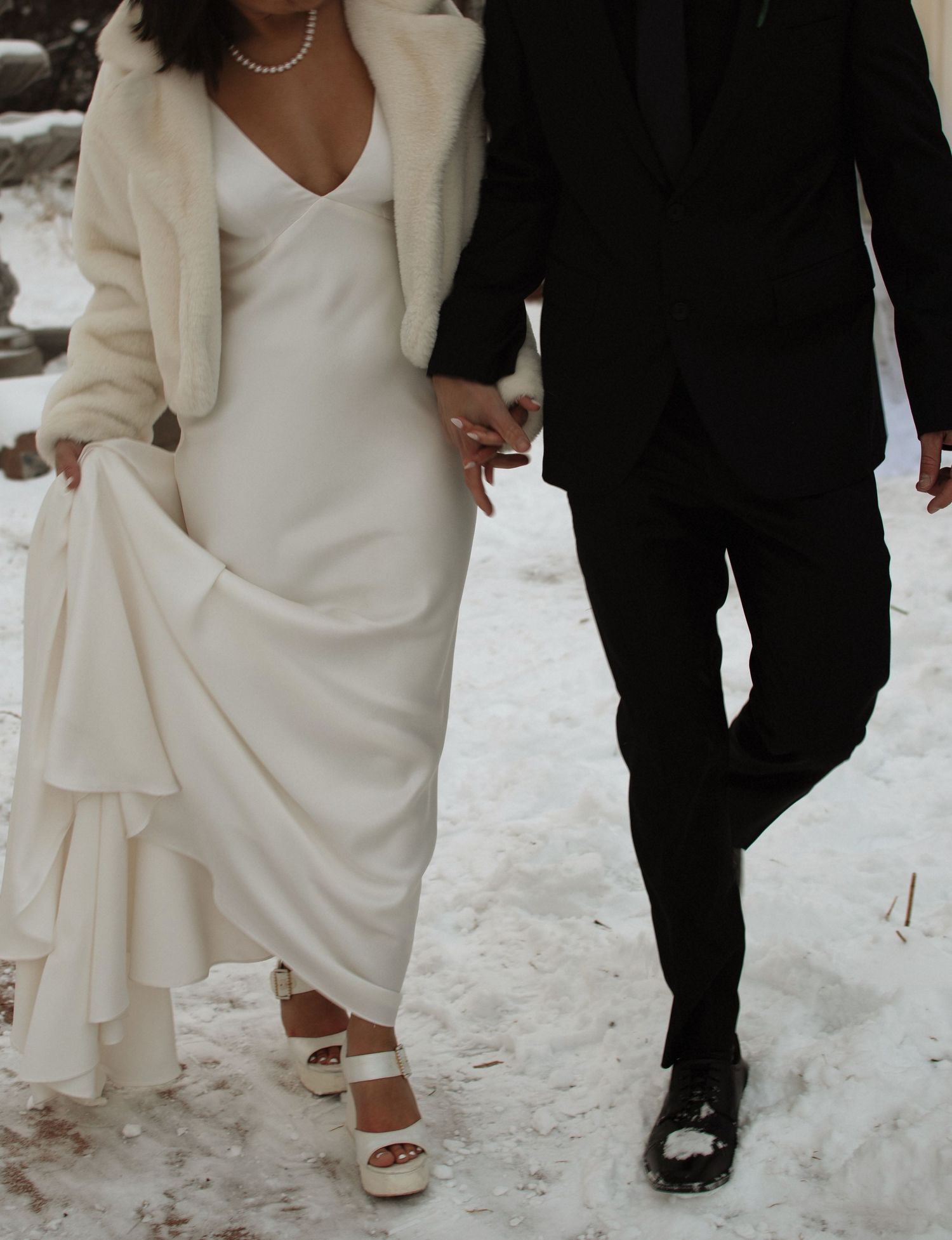 Bride in white slip dress and fur shawl holds groom's hand walking through snow in winter wedding setting.