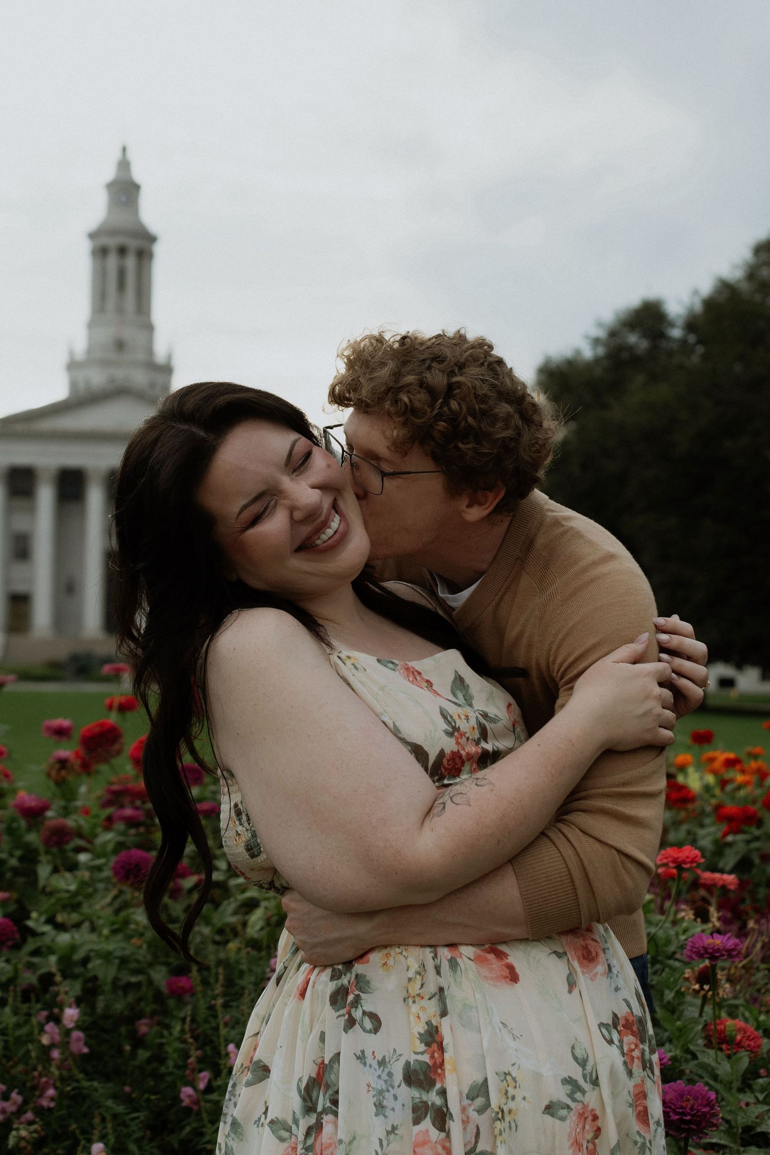 Couple embracing in a flower garden, woman laughing as man kisses her cheek, historic building in background.