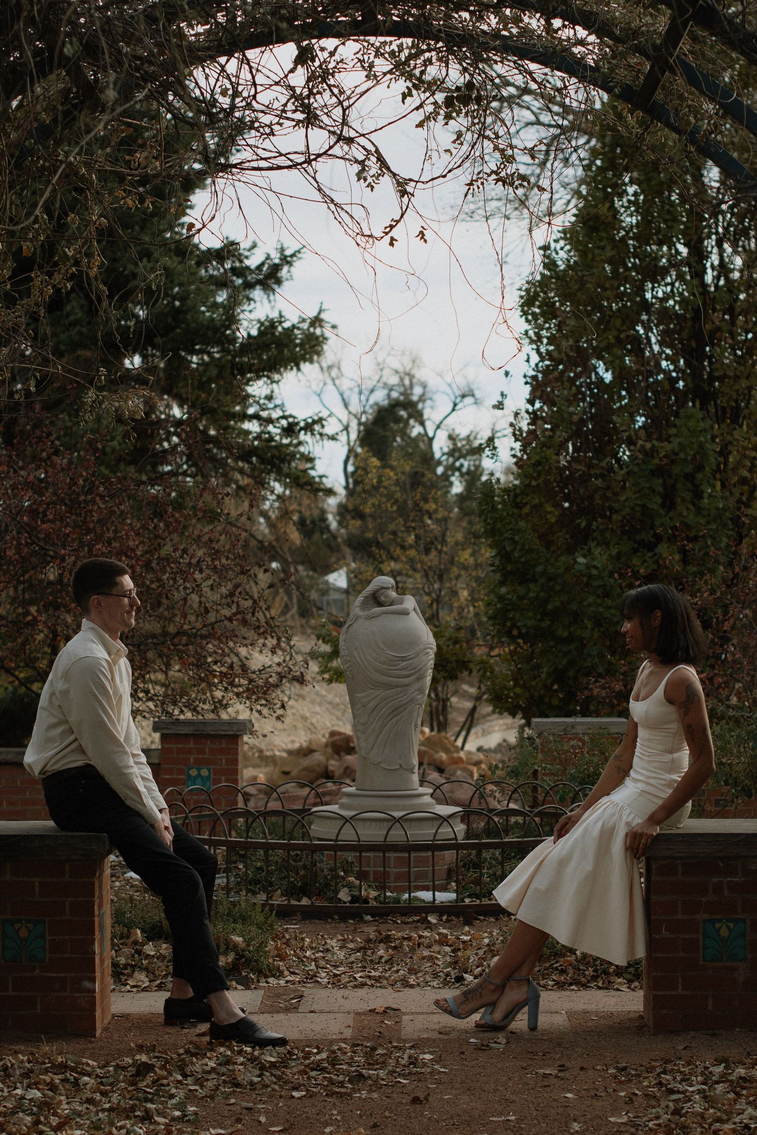 Couple in elegant attire sitting apart under garden arch with statue backdrop.
