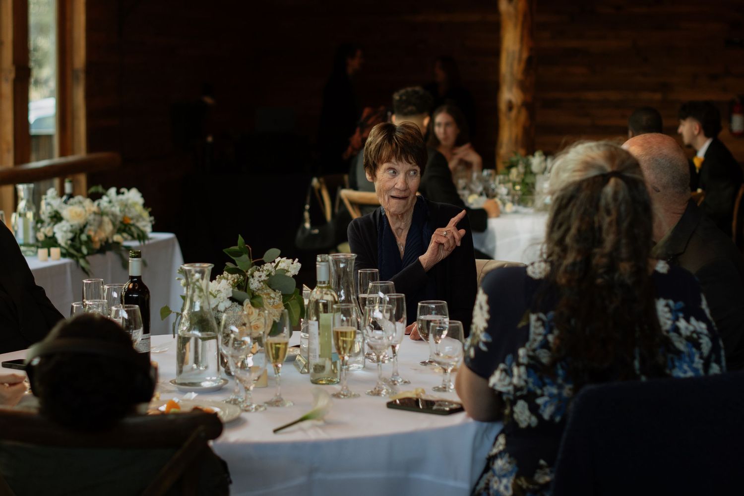 Guests gathered around elegant white tables at a rustic wedding reception in a log cabin venue.