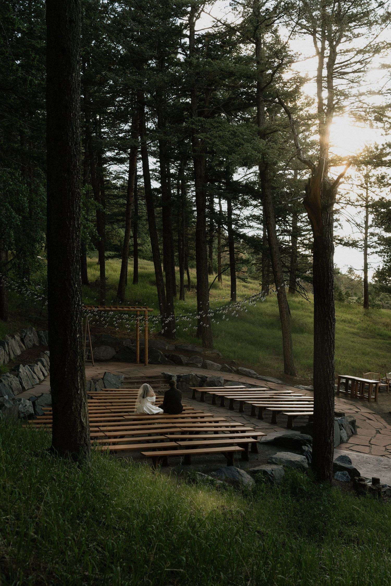 Couple sits together on wooden benches in a sunlit outdoor amphitheater surrounded by tall trees at golden hour.