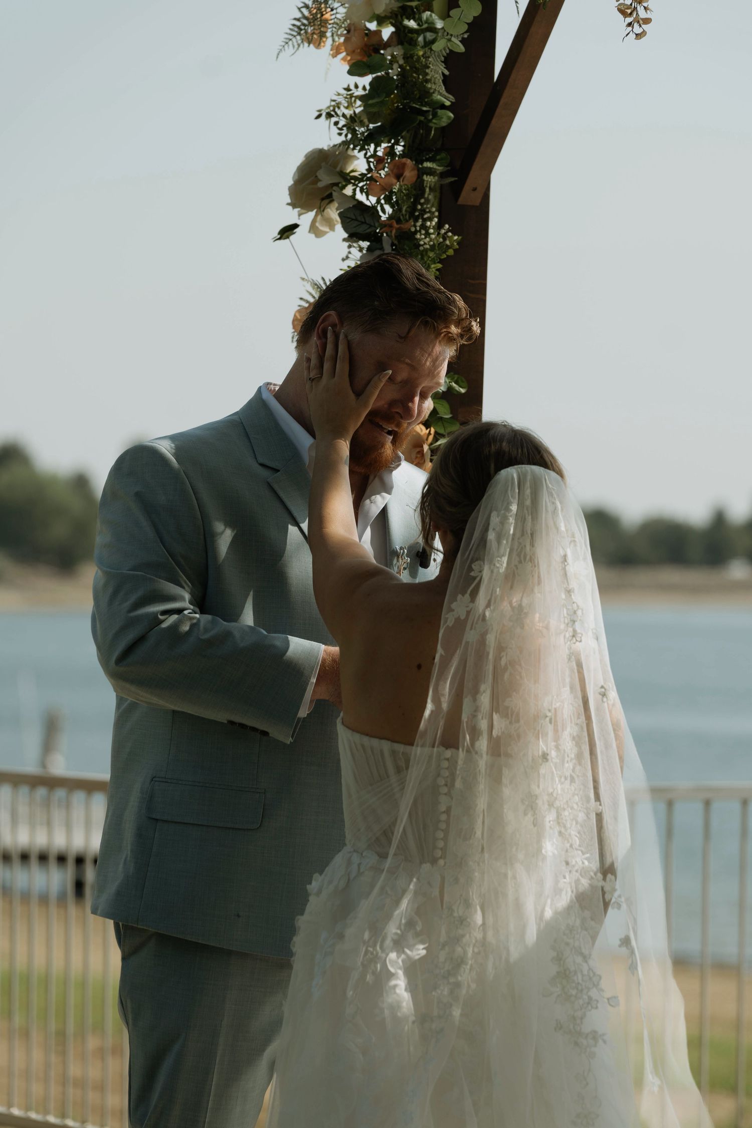 Bride tenderly touches groom's face during outdoor waterside wedding ceremony under floral arch, groom in sage suit, bride in lace veil.