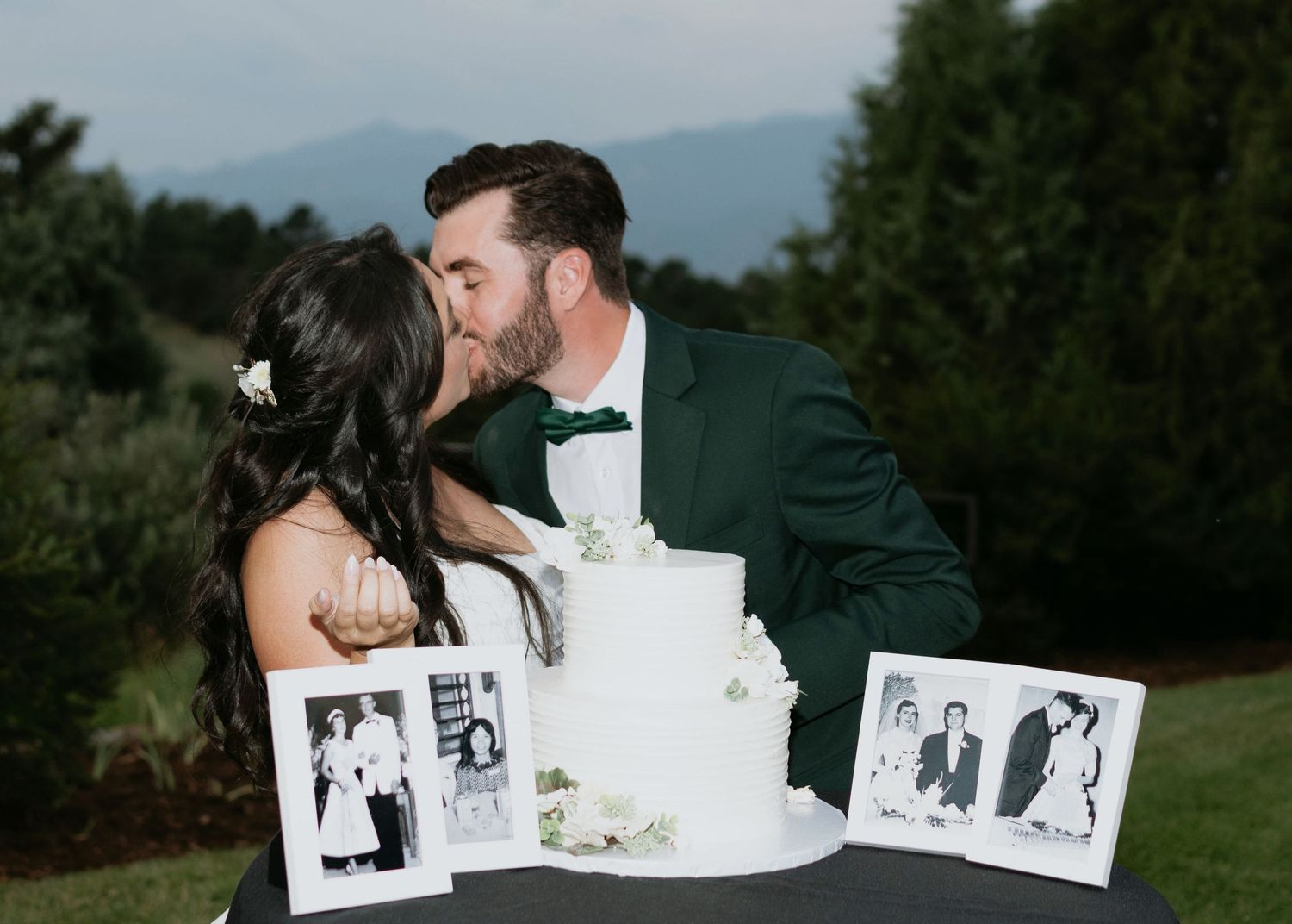 flash photos of bride and groom kissing over wedding cake after cake cutting, mountain views and green grass