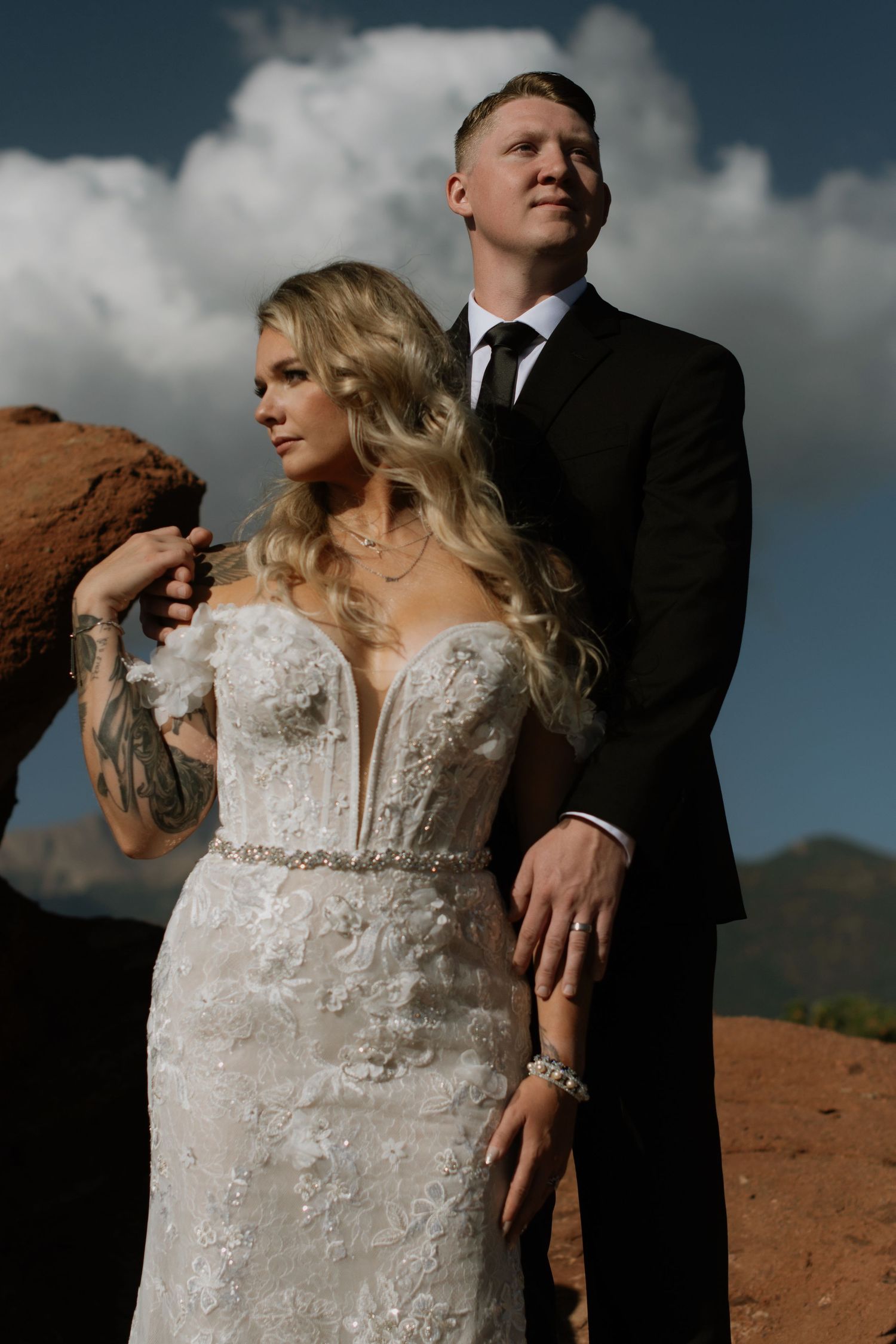Bride in floral lace gown and groom in black suit pose among red rocks under dramatic cloudy sky.