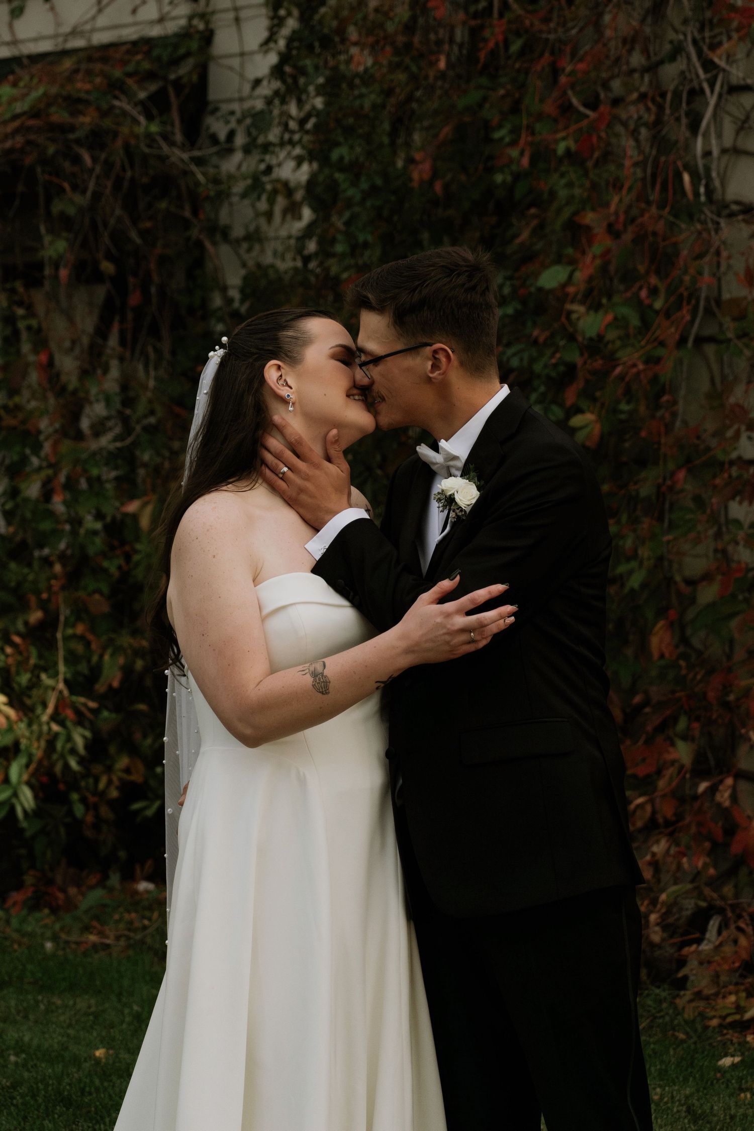 A bride and groom share a romantic kiss outdoors in a garden setting, dressed in wedding attire.