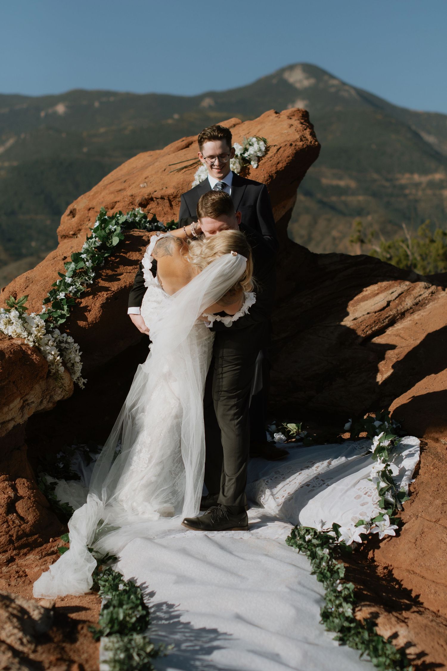 Bride and groom share first kiss at outdoor wedding ceremony among red rocks with mountain backdrop in Colorado.