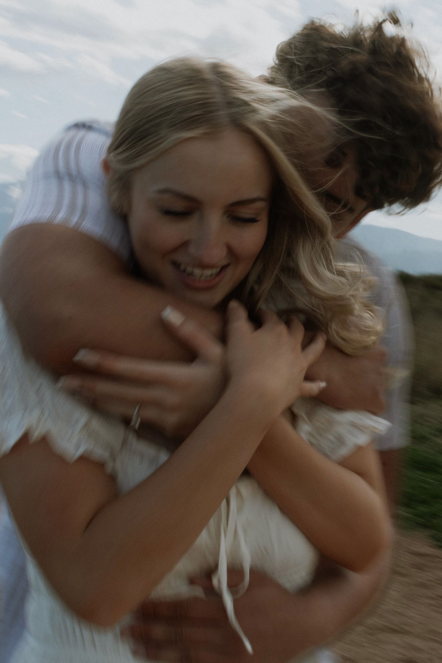 A joyful blonde woman smiles as a curly-haired man embraces her from behind outdoors.