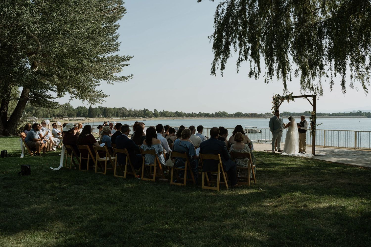 Outdoor wedding ceremony by a lake with guests seated on wooden chairs facing couple at a wooden arch altar.