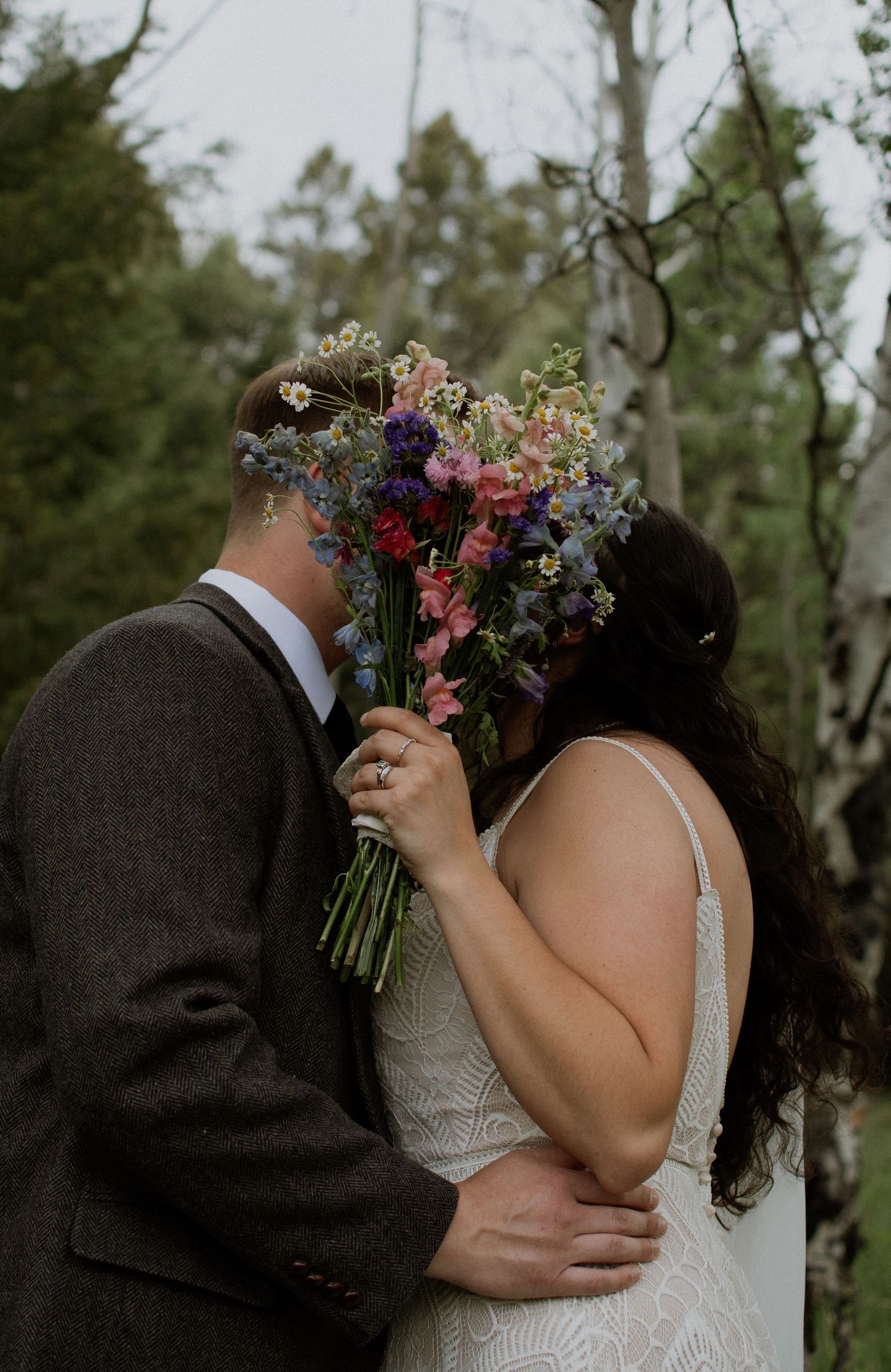 A couple shares a kiss hidden behind a colorful wildflower bouquet in a forest wedding setting.