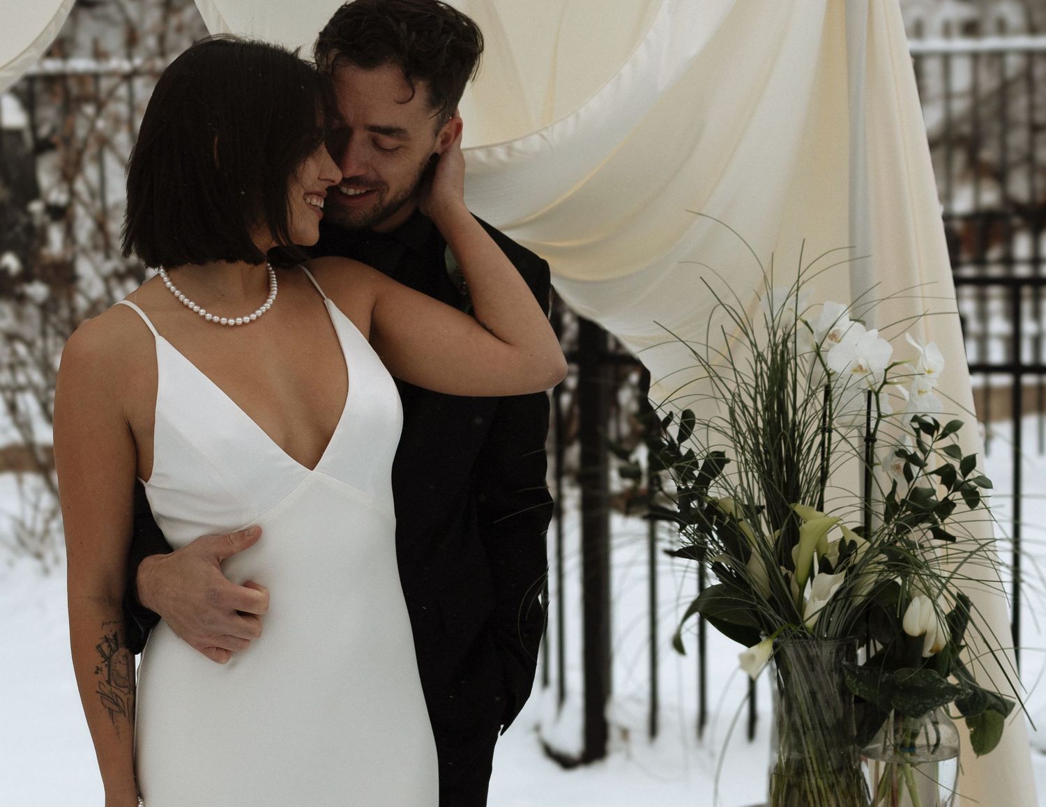 Couple embracing at outdoor winter wedding ceremony with white drape backdrop and floral arrangement in snow.