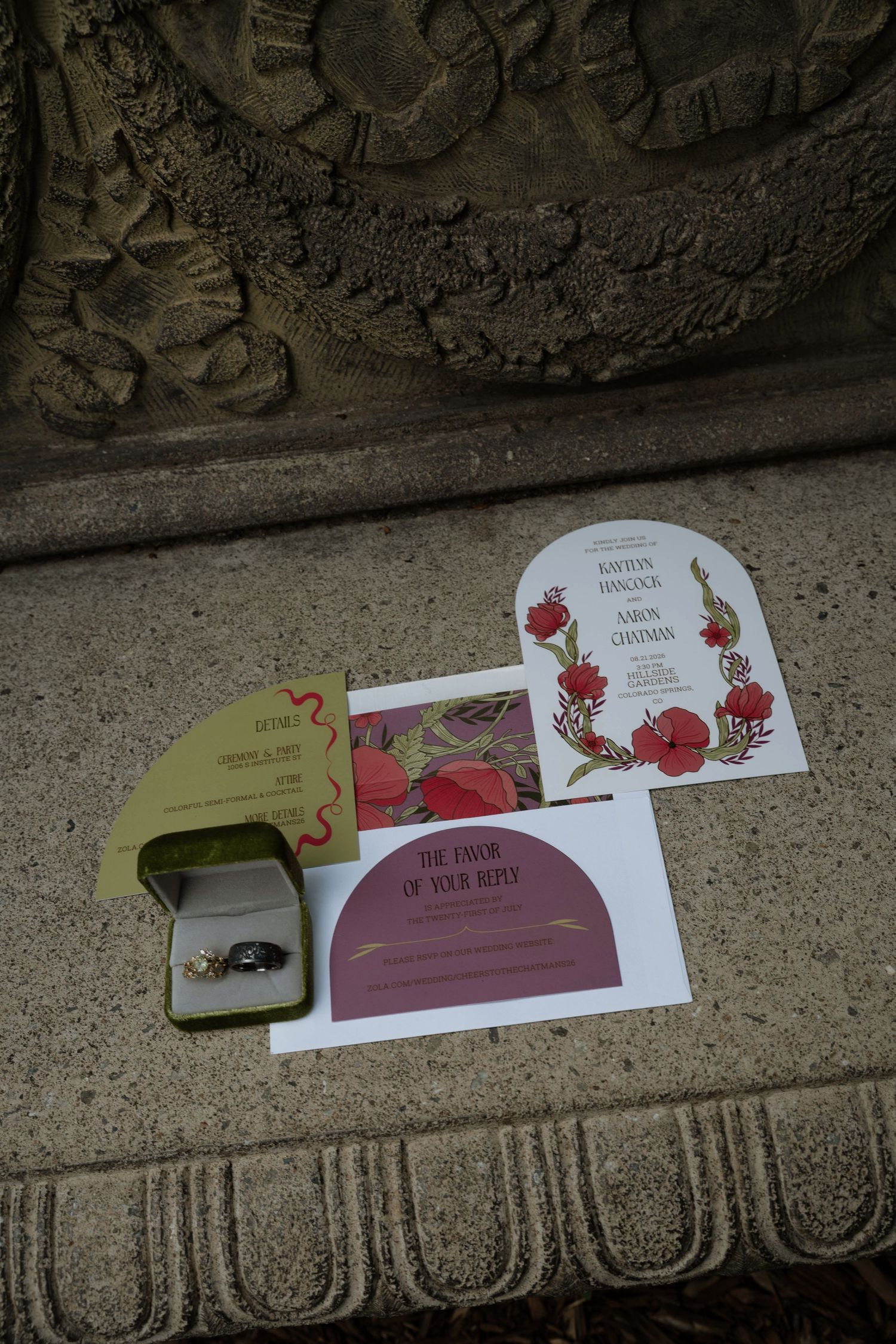 Wedding invitation suite with floral red poppy designs, RSVP card, and diamond ring in velvet box on stone surface.