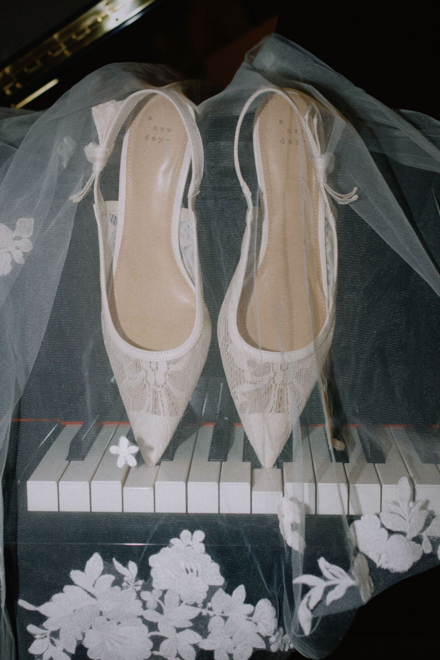 White lace bridal heels with ankle straps resting on piano keys beneath a floral wedding veil.