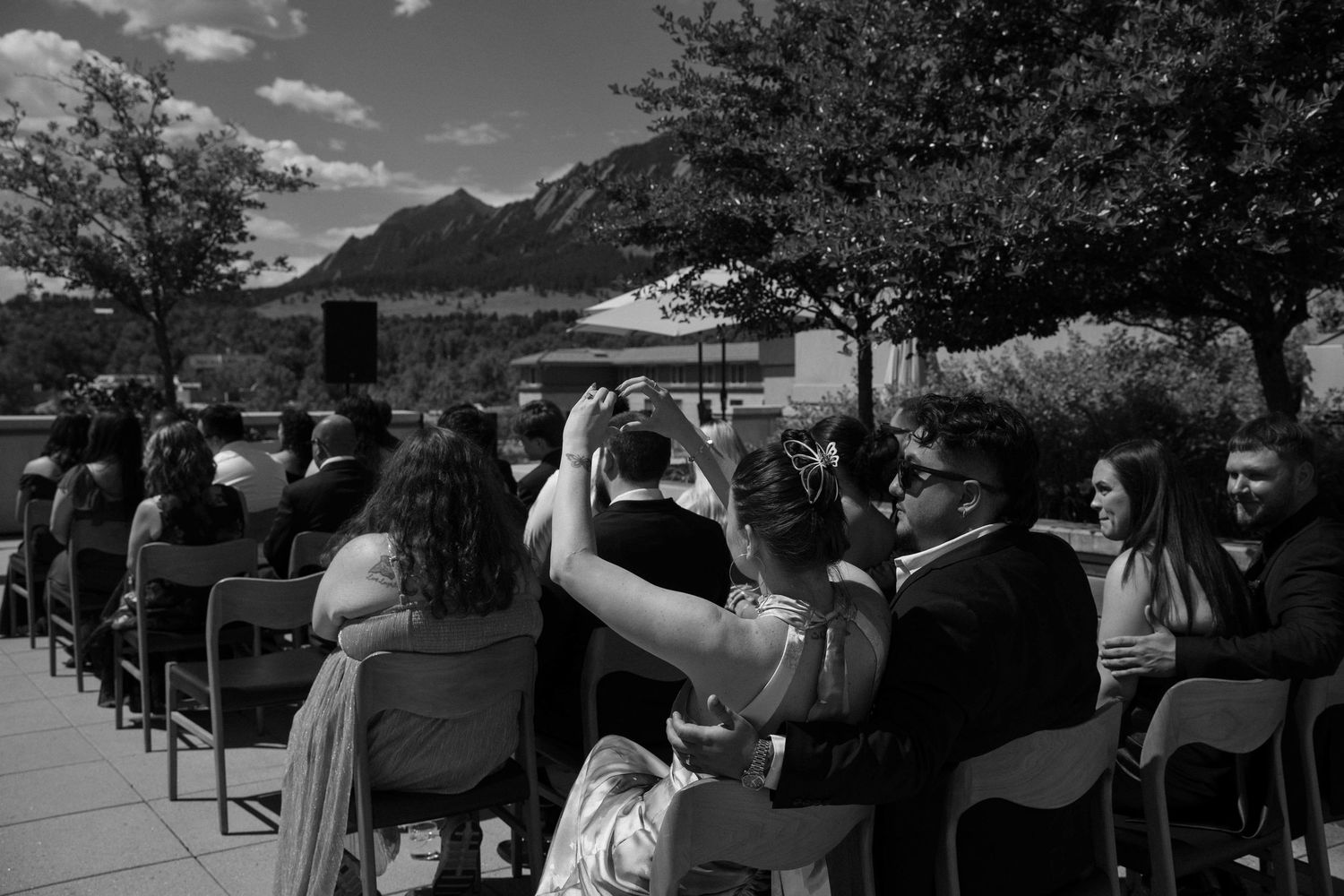 Black and white outdoor wedding ceremony with guests seated, mountains and trees in background.