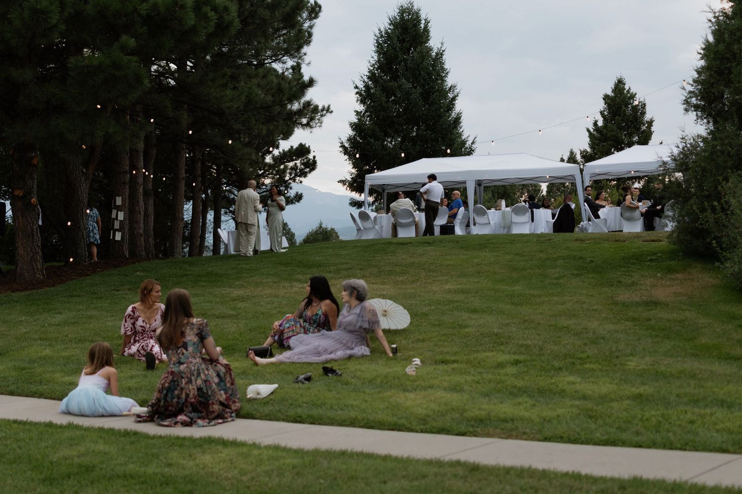 Outdoor wedding reception on a green lawn with guests relaxing on grass and a white tent venue in the background.