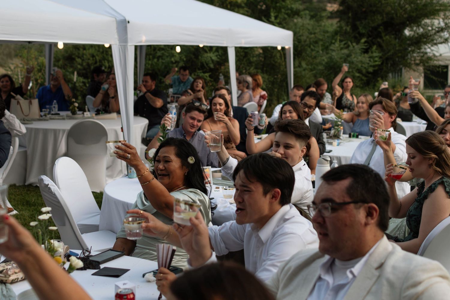 family and friends, guests cheers at all white tables with tent and string lights in the trees
