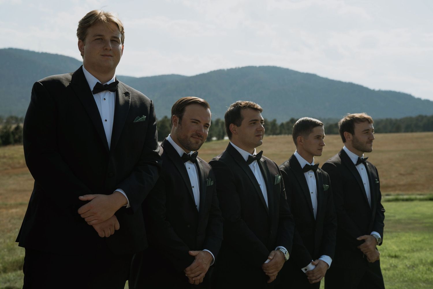 Five groomsmen in black tuxedos stand outdoors at a wedding with mountain scenery in the background.