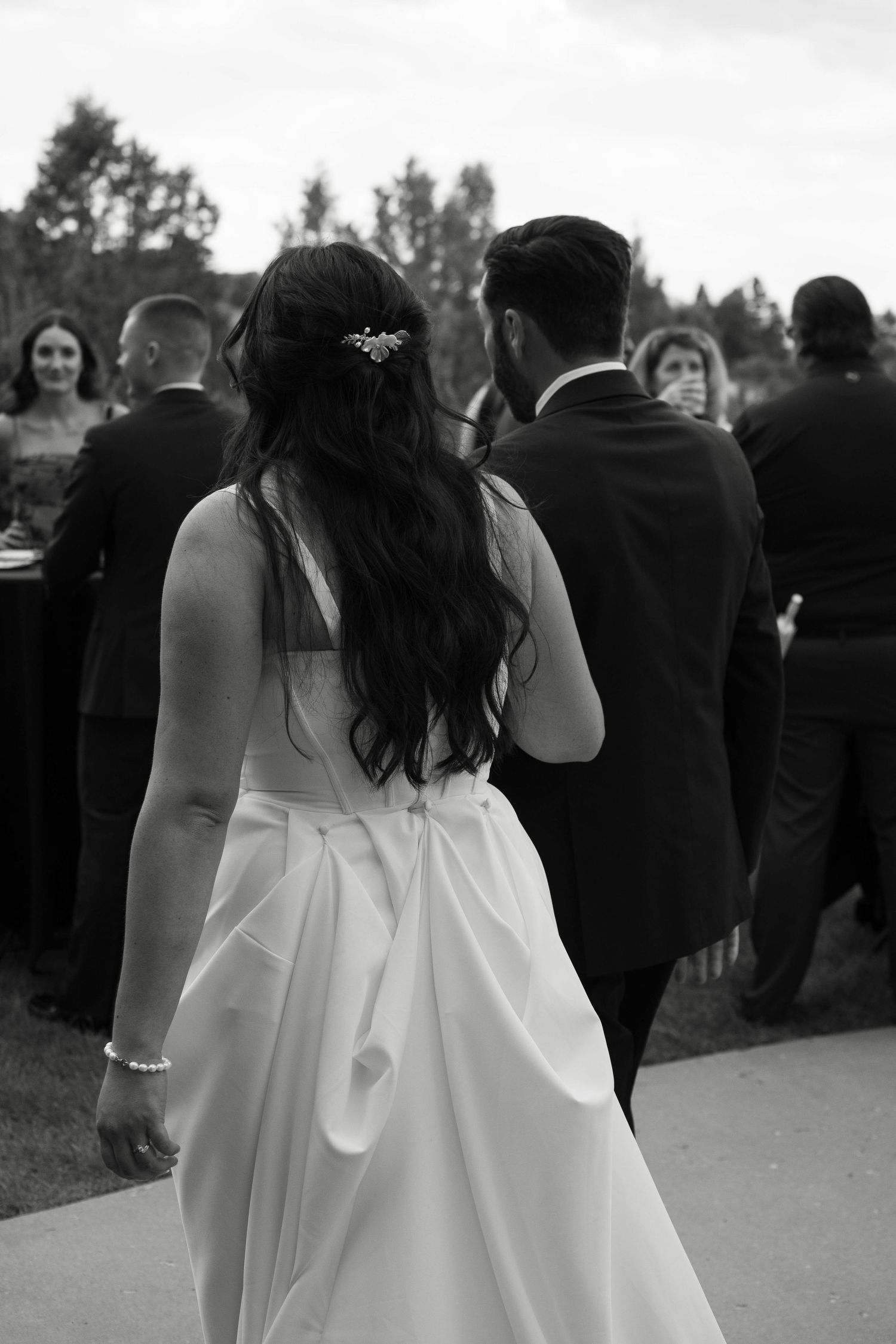 Black and white photo of bride in white gown and groom in tuxedo walking toward wedding guests outdoors.