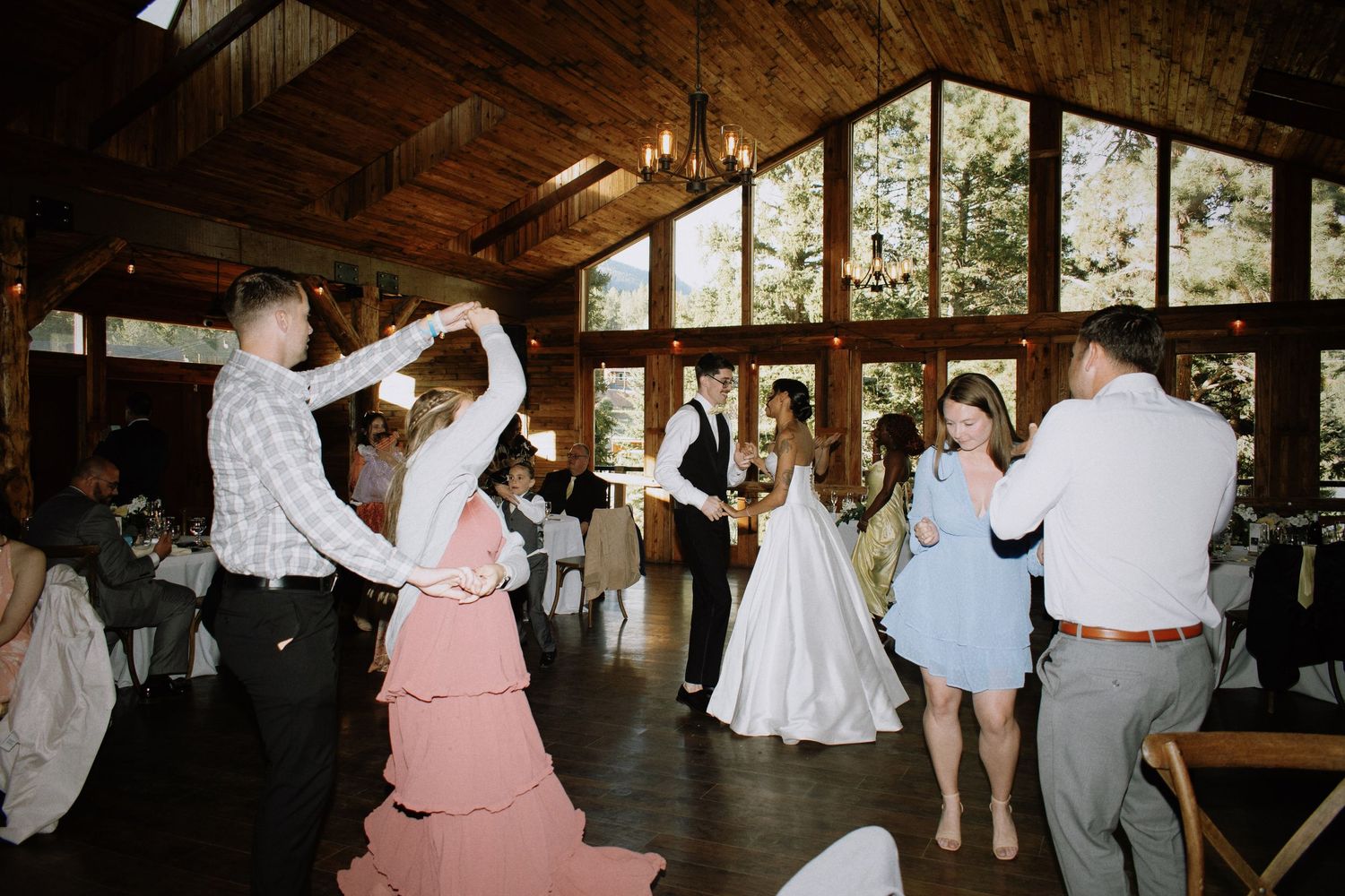 Wedding guests dancing joyfully in a rustic wooden venue with high ceilings and chandeliers.
