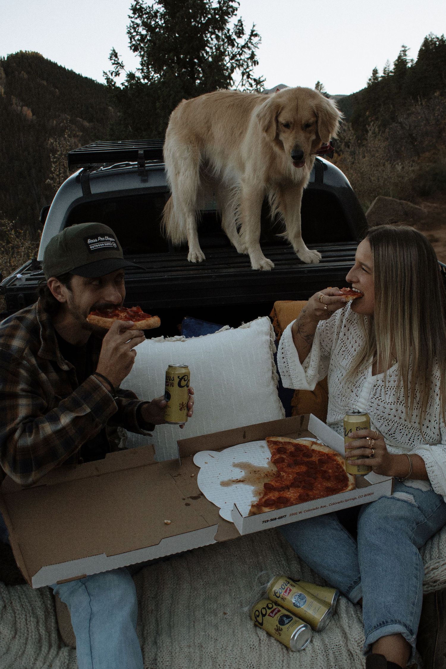 Couple eating pepperoni pizza and drinking beer in truck bed with golden retriever dog, mountains in background.