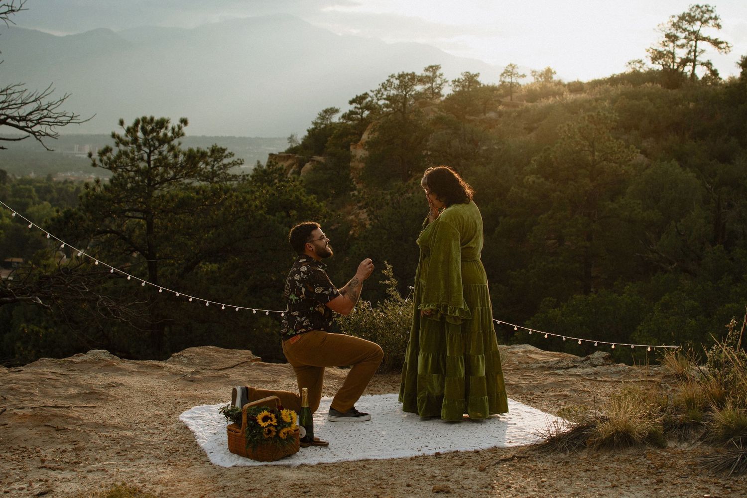 Man proposes on one knee to woman in green dress on rocky overlook at sunset with string lights and trees.