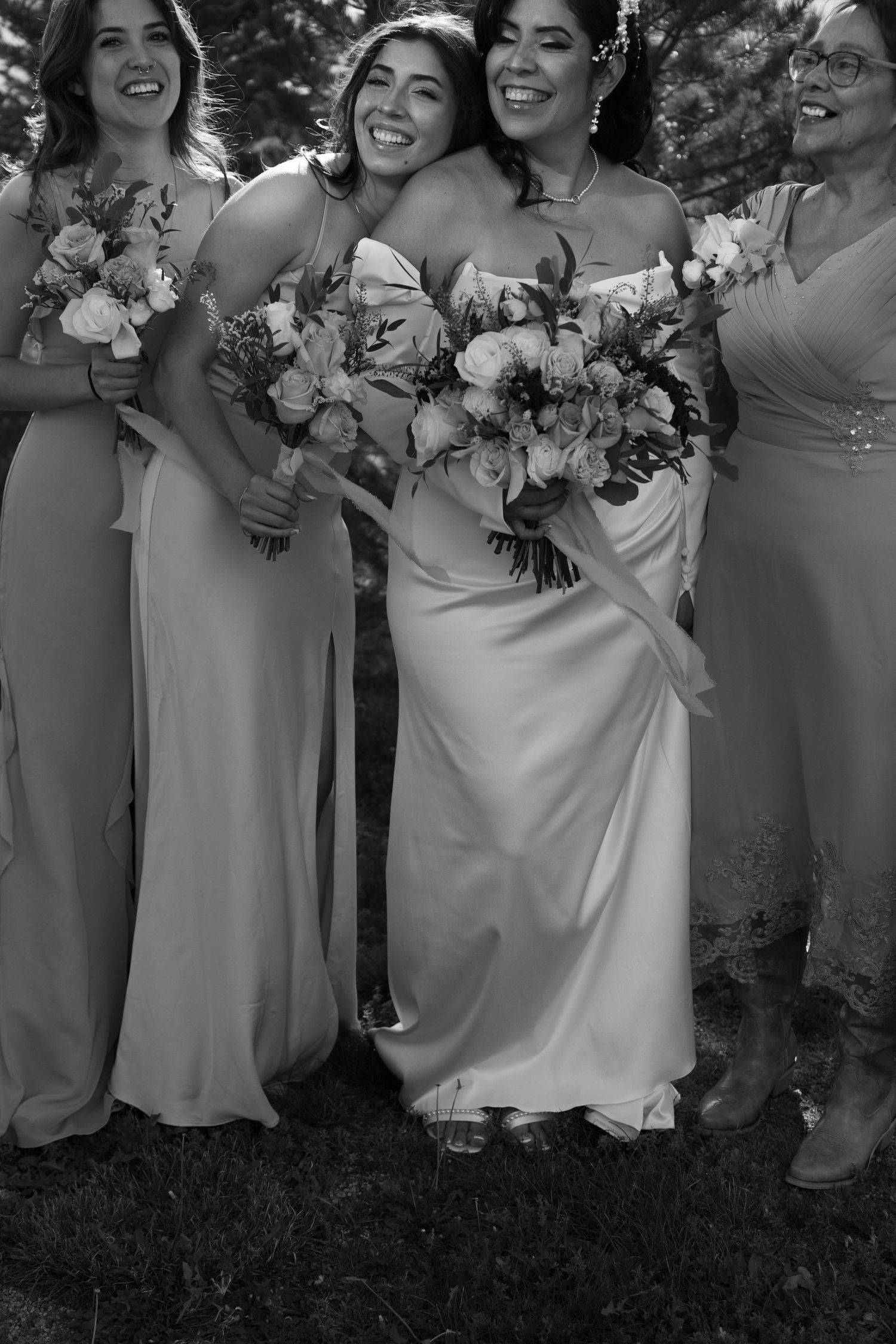 Black and white photo of a smiling bride in a white gown hugging her bridesmaids while holding floral bouquets.