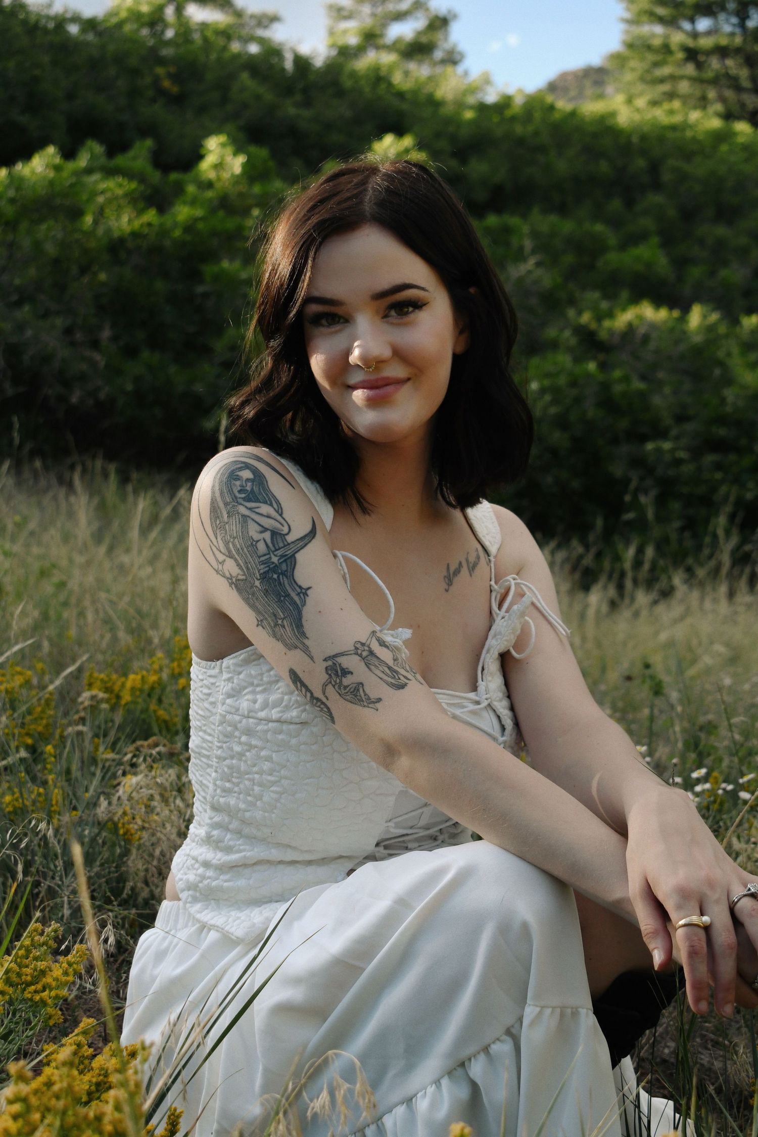 Smiling brunette woman with tattoos wearing a white sundress sitting in a wildflower meadow outdoors.