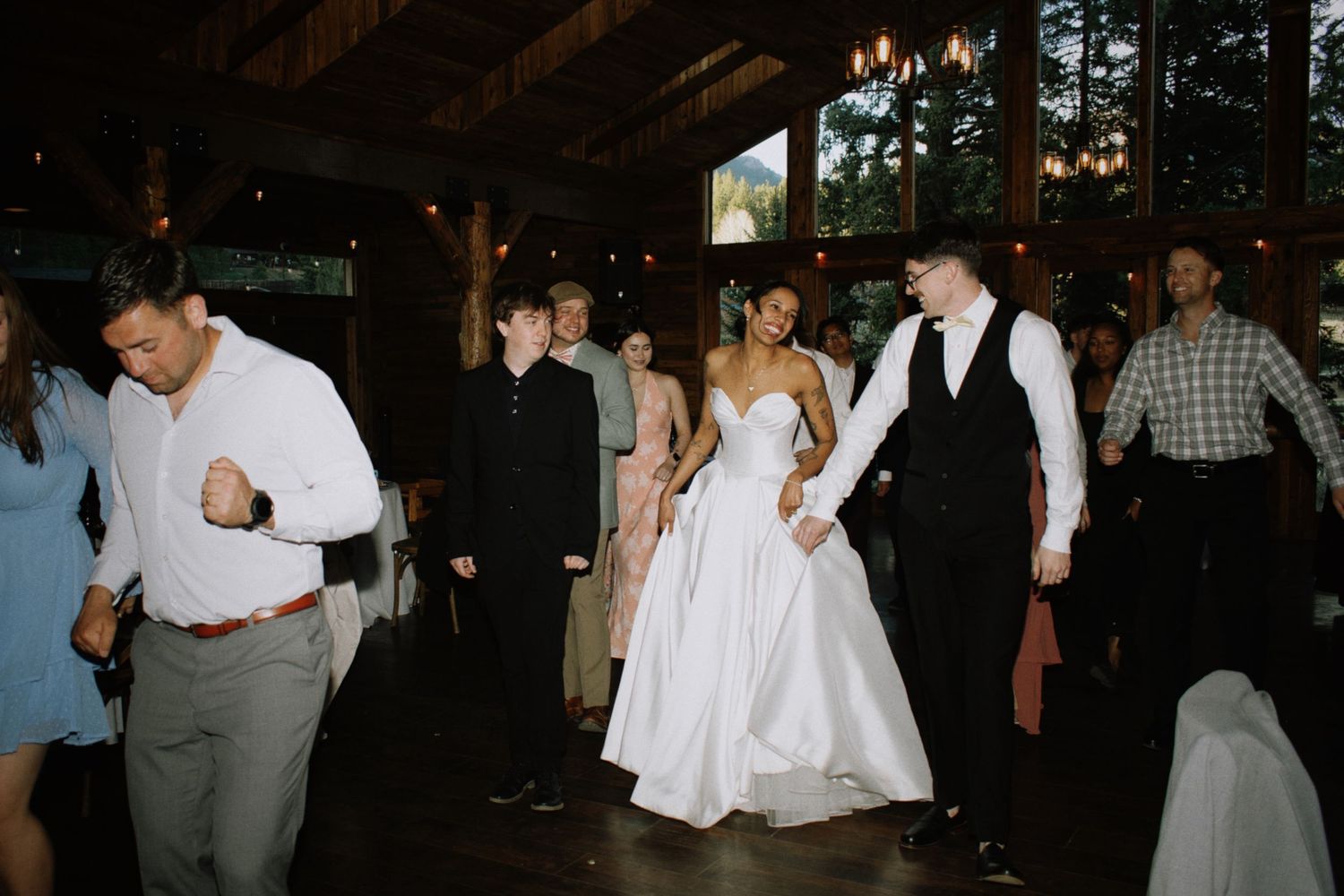 Bride in white ballgown dancing with guests at lively rustic barn wedding reception with chandelier overhead.