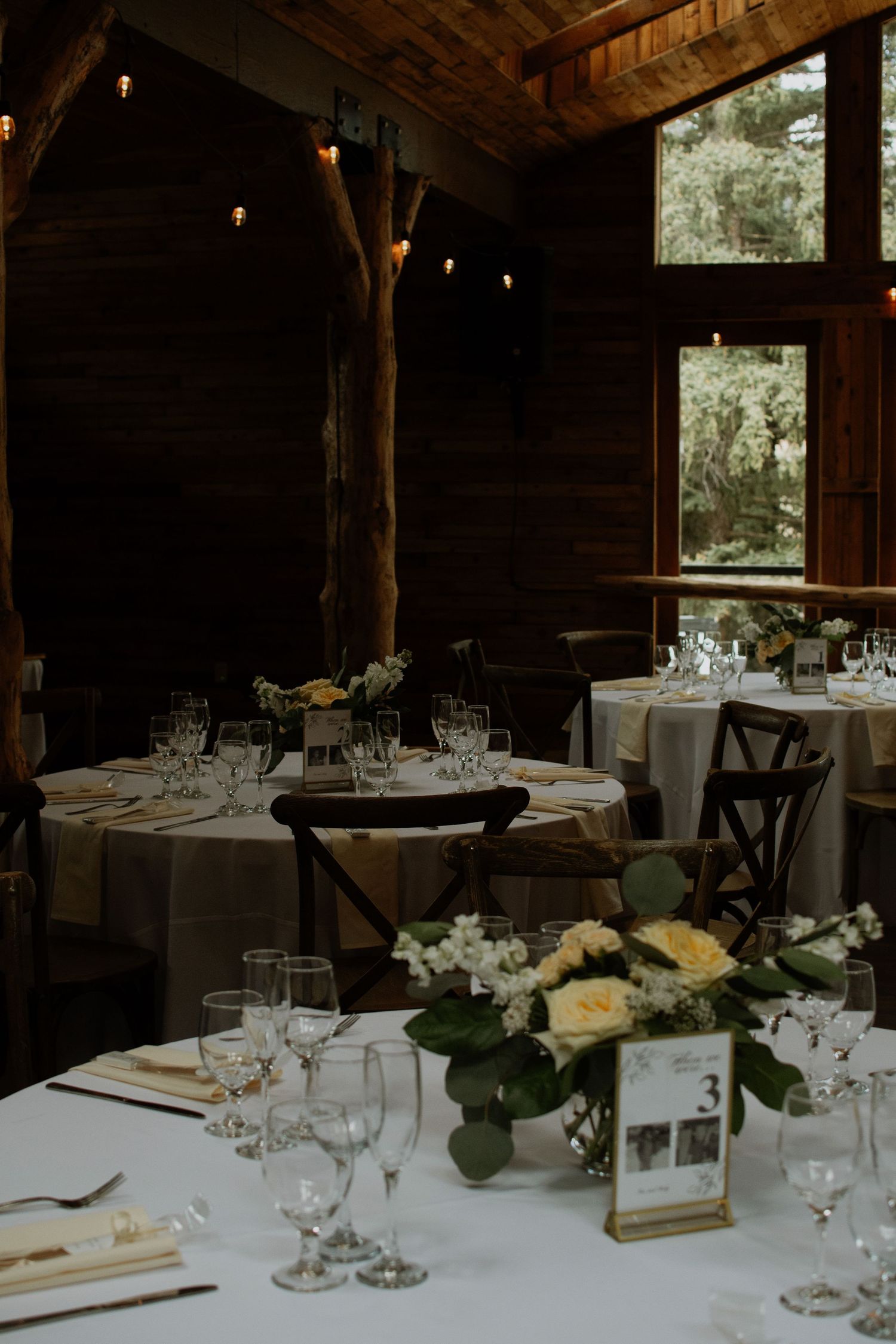 Elegant rustic barn wedding reception with round tables, white floral centerpieces, string lights, and cross-back chairs.