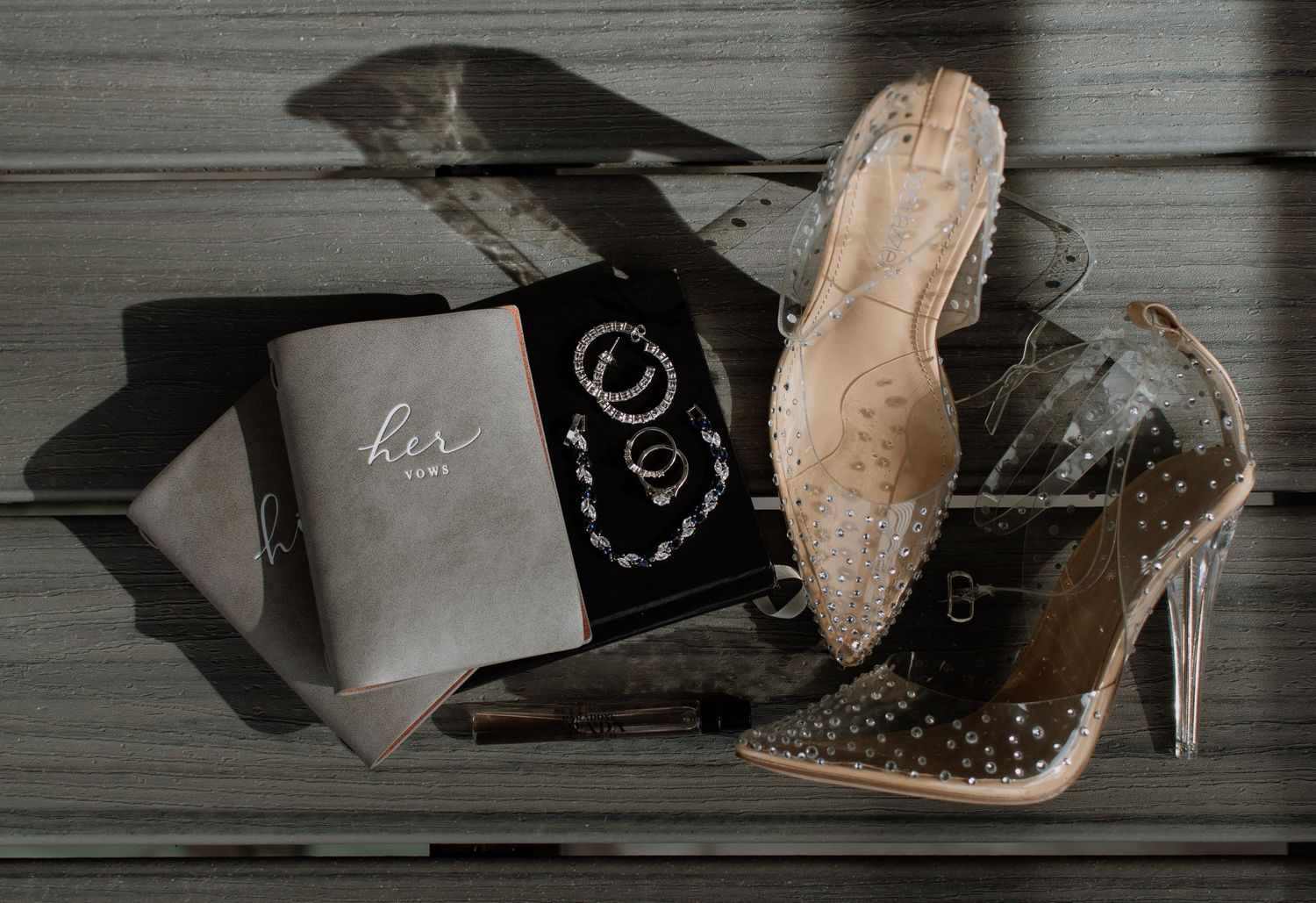 Elegant wedding flat lay with clear studded heels, gold rings, and a gray vow book on a wooden surface.