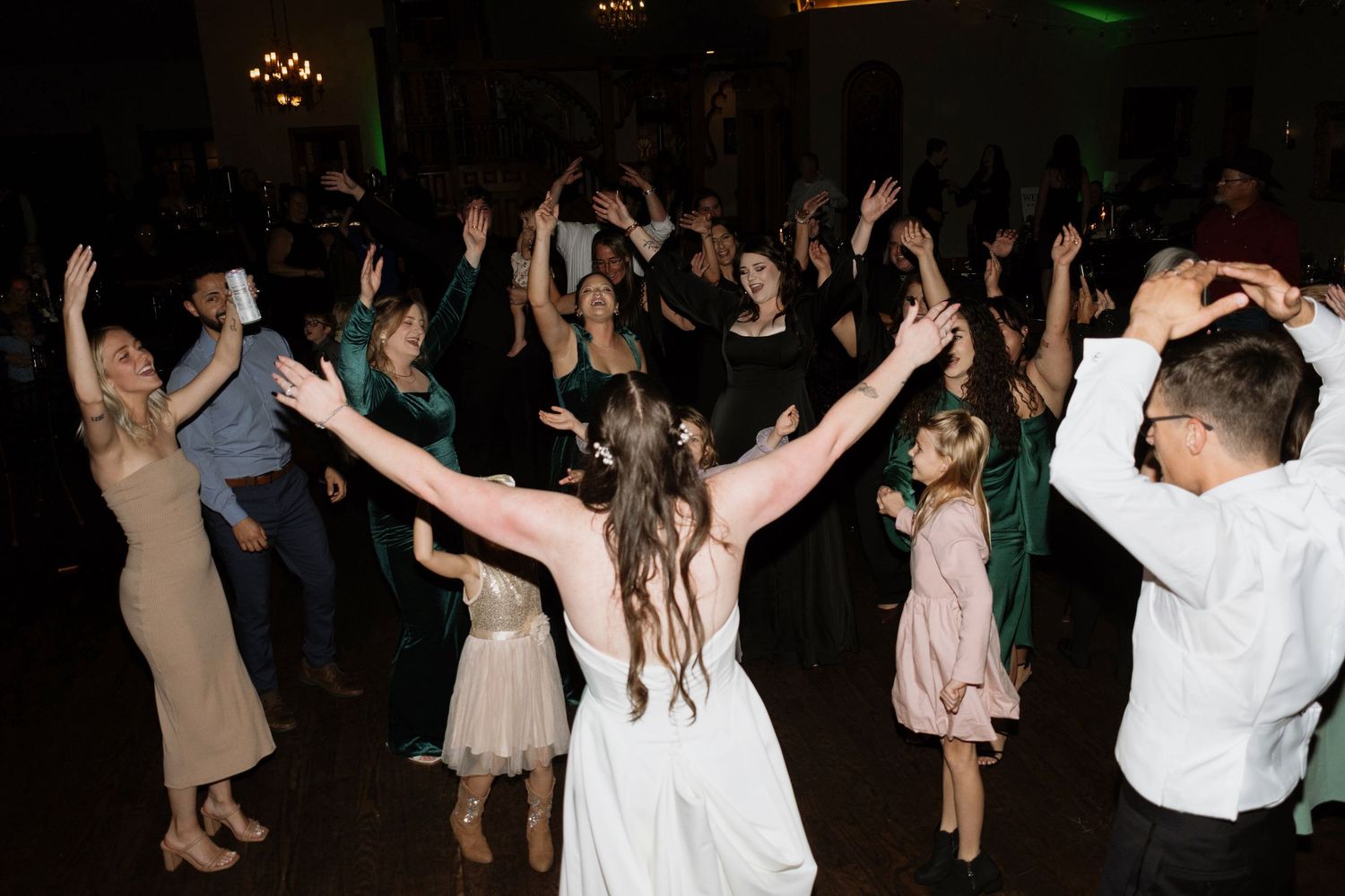 Bride and guests dancing with arms raised at a lively wedding reception.