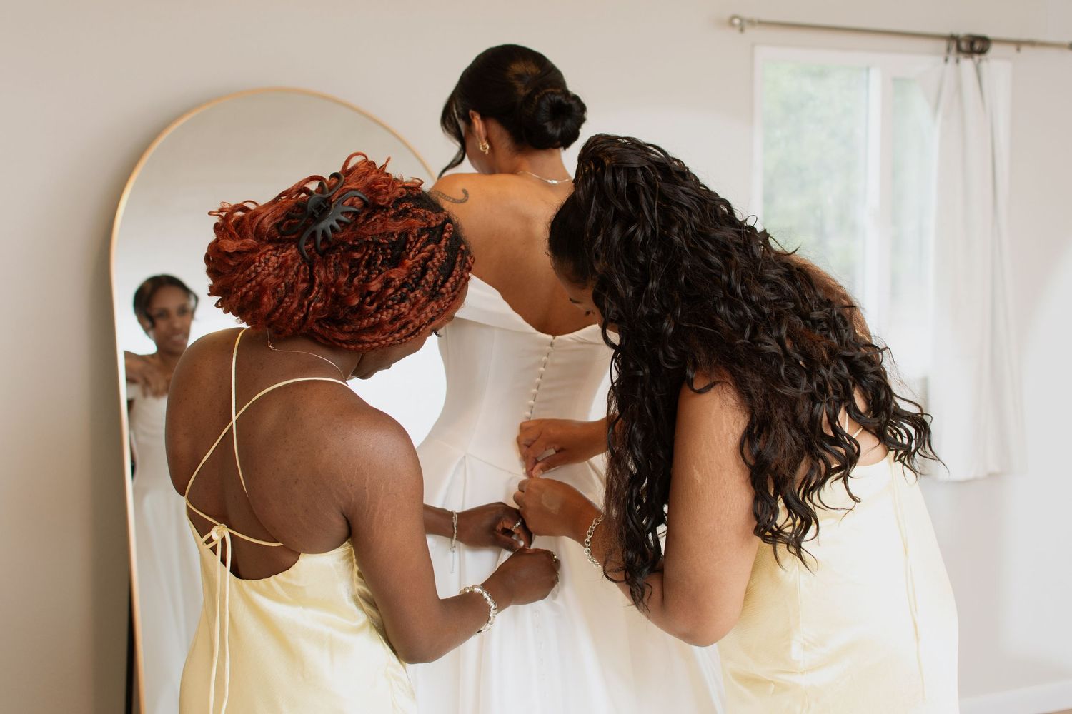 Two bridesmaids in yellow dresses helping a bride button her white wedding gown in front of a mirror.