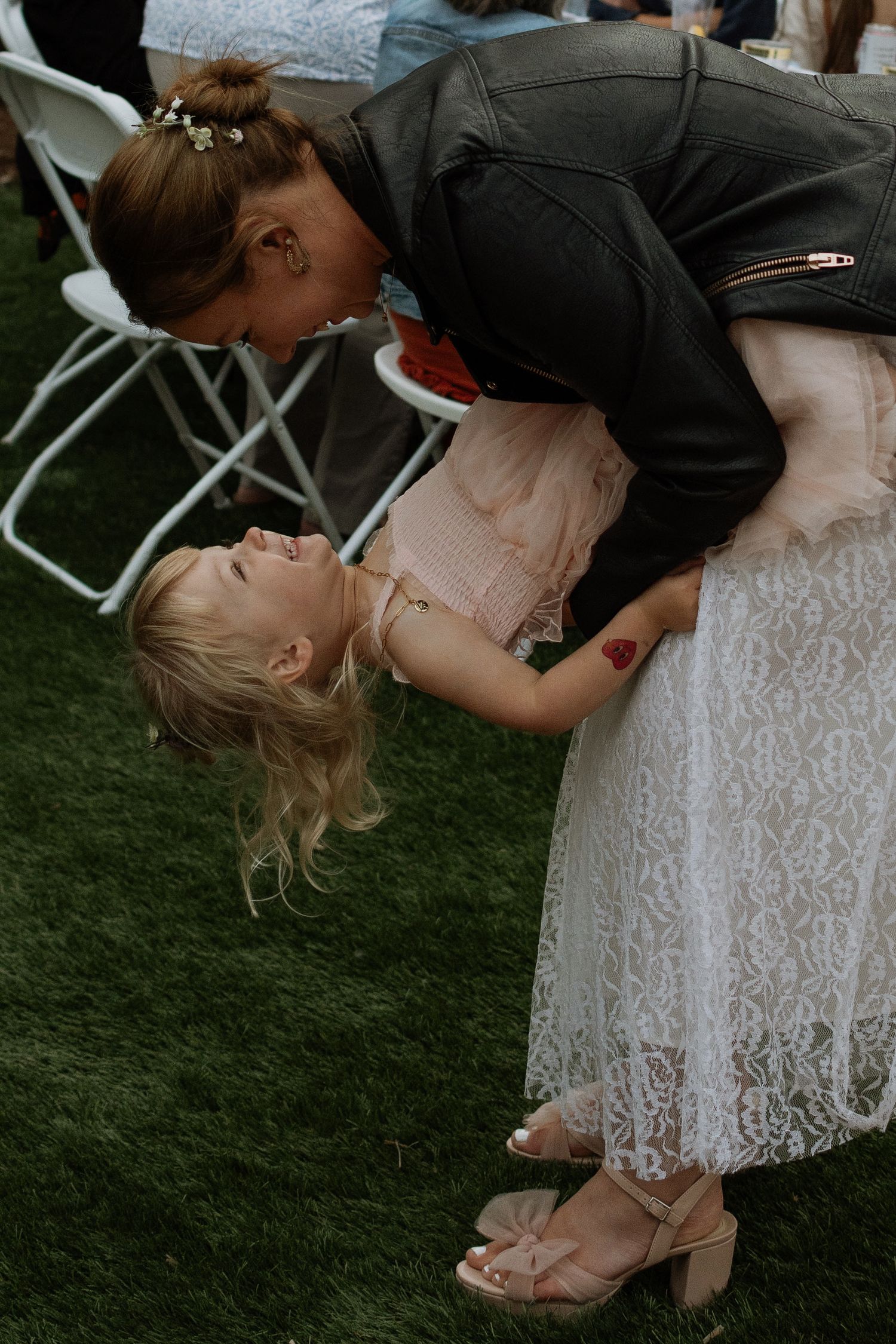 Woman in leather jacket playfully dipping laughing blonde girl in lace dress at outdoor wedding reception.