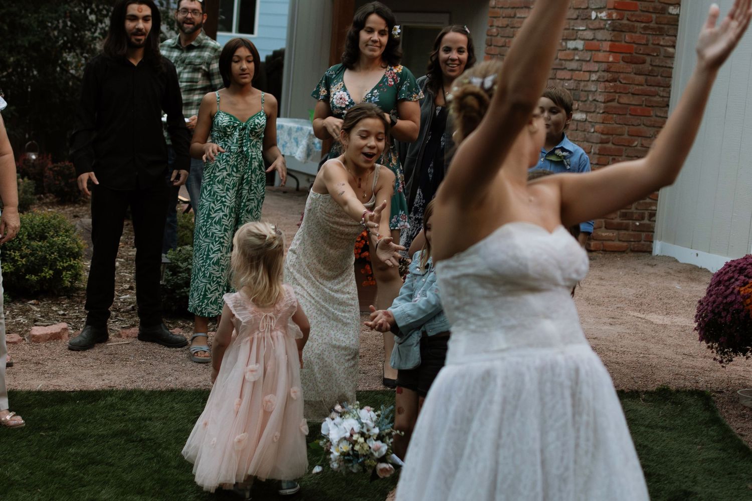 Bride tossing bouquet to excited wedding guests including a flower girl in a pink dress outdoors.