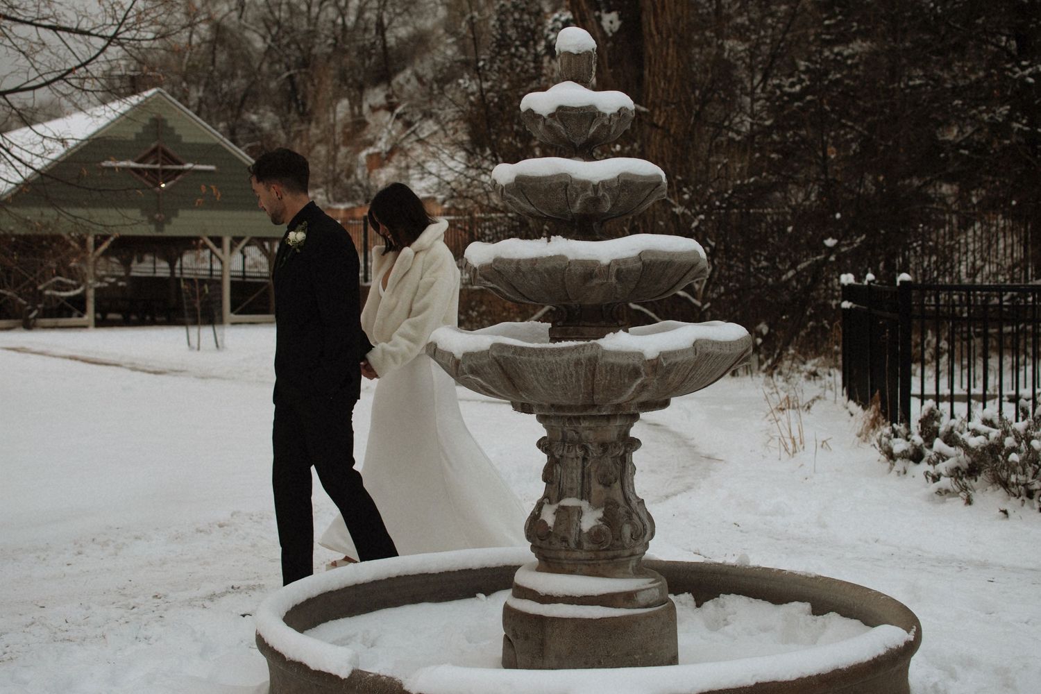 Bride and groom stand by a snow-covered stone fountain in a winter wedding outdoor setting.