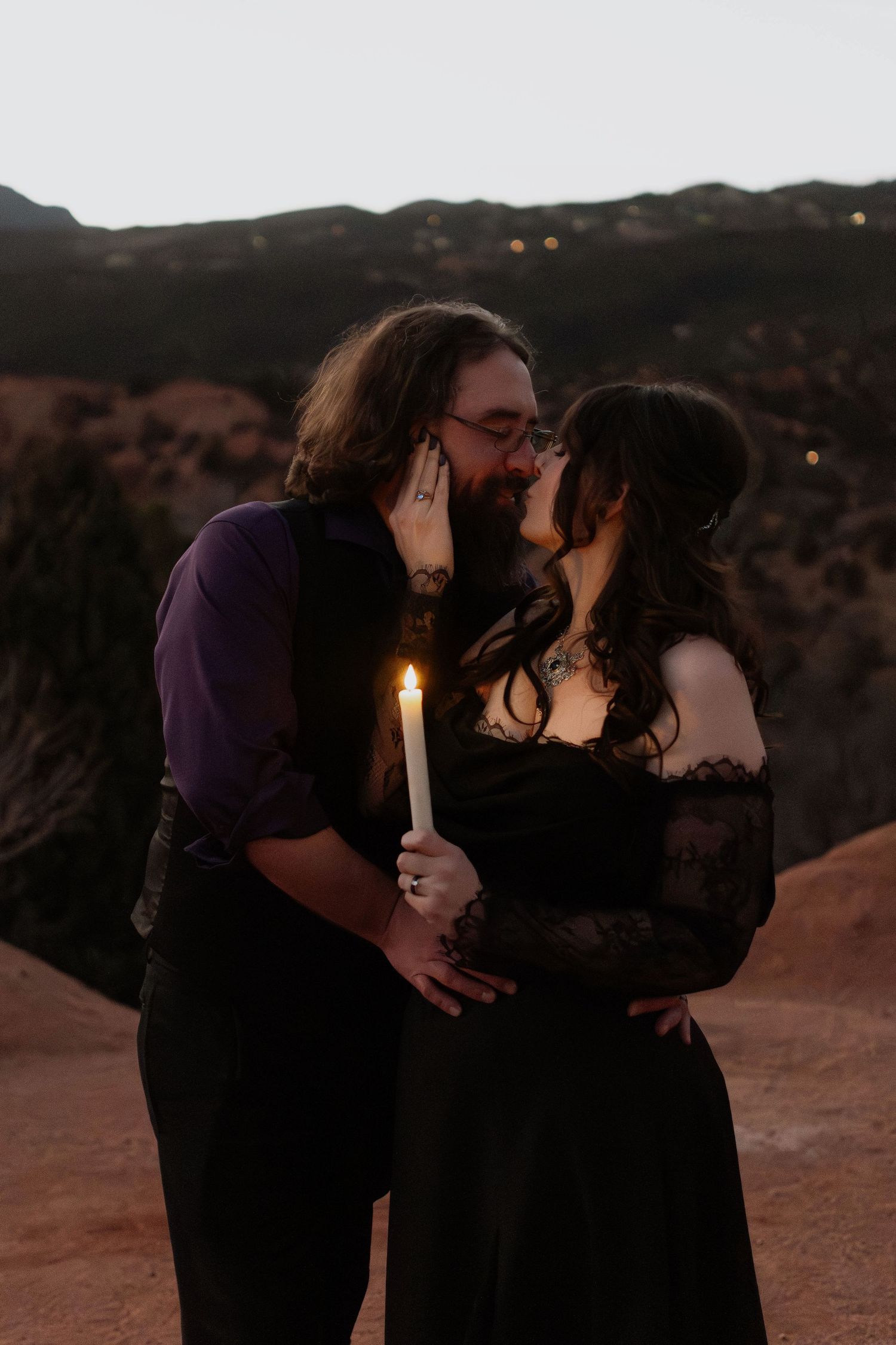 Couple sharing a romantic kiss at dusk in a mountain landscape, woman holding a lit white candle, both dressed in elegant black attire.