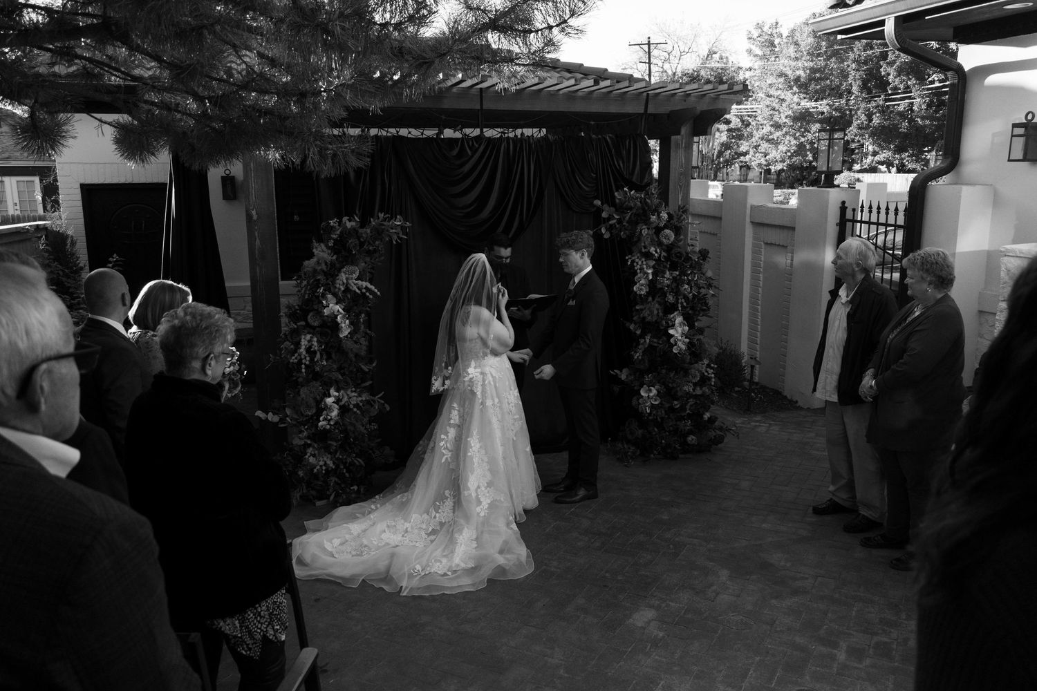 Black and white photo of bride and groom exchanging vows at an outdoor wedding ceremony with guests.