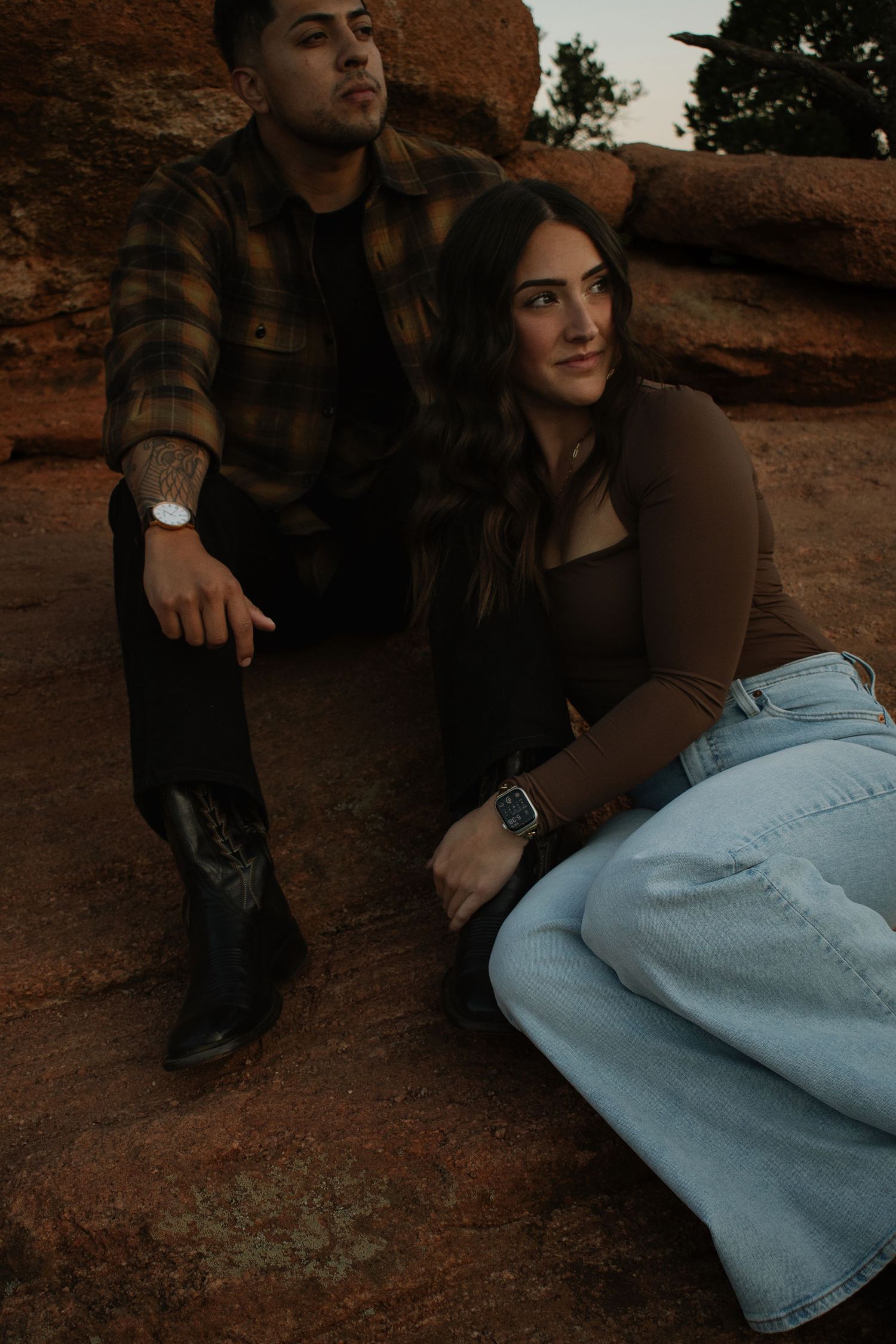 A couple sits together on red rock formations, wearing earth tones, sharing a quiet moment at golden hour.