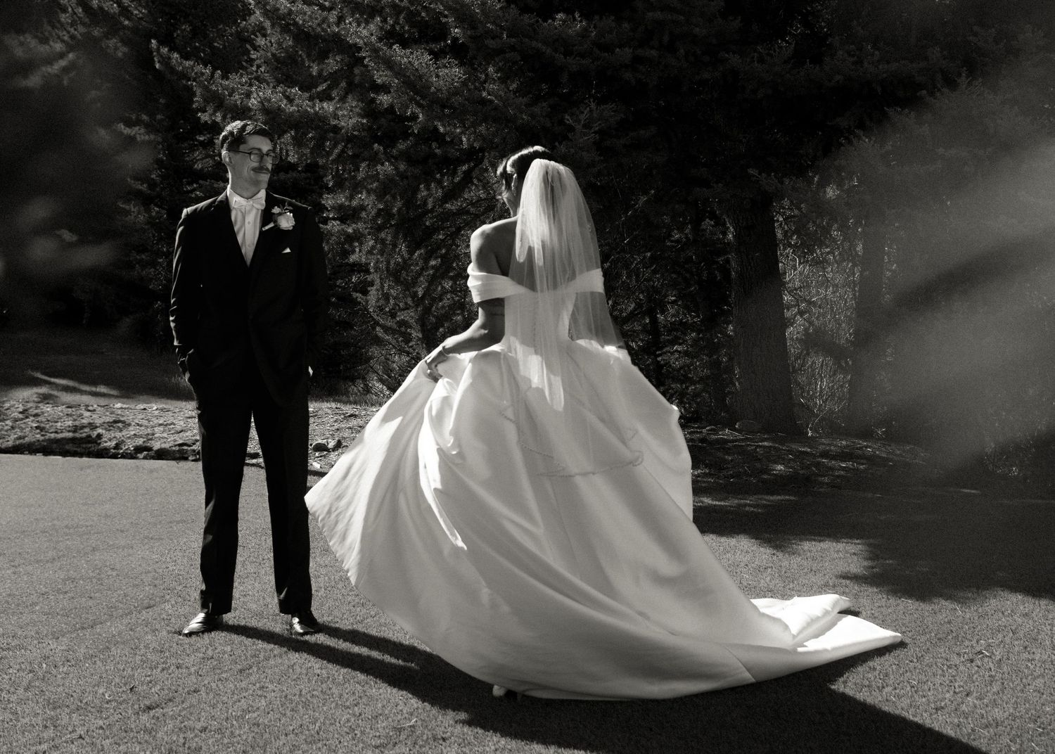 black and white photo of groom admiring bride tailing in her classic off the shoulder ball gown and veil. mixed race Colorado forest wedding