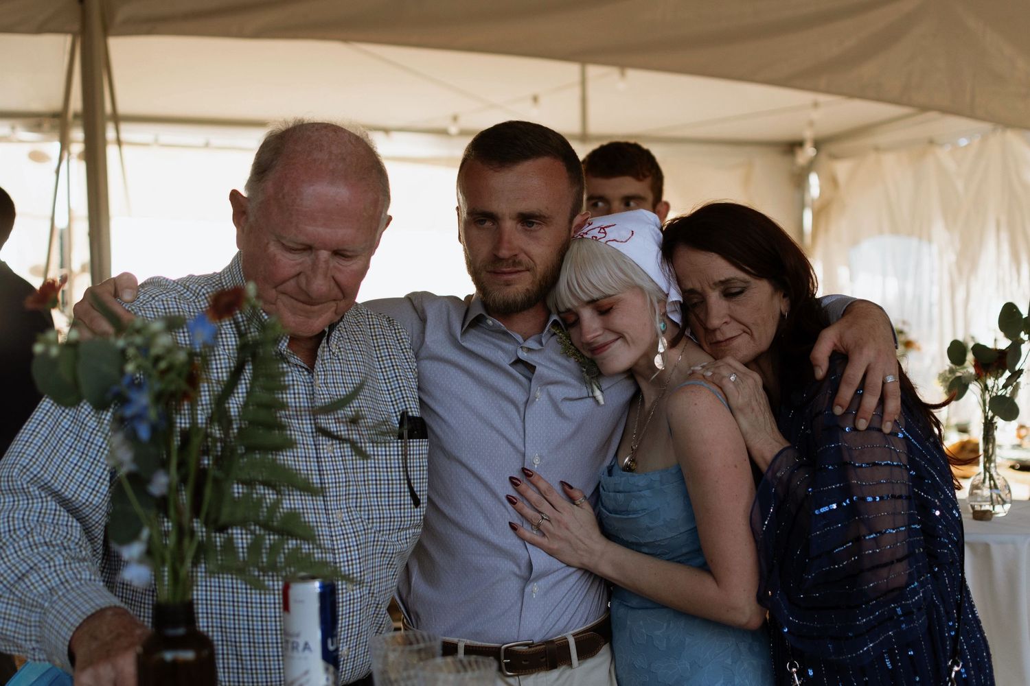 Family group hug at a wedding reception under a tent, sharing an emotional moment together.