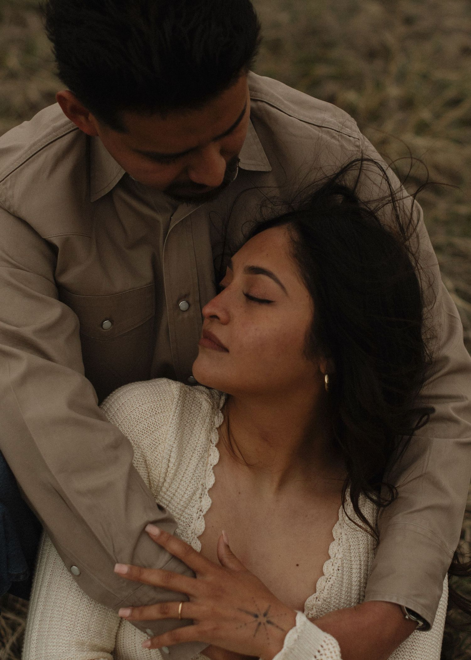 A couple shares an intimate embrace outdoors, the woman in white lace resting against a man in a beige shirt.