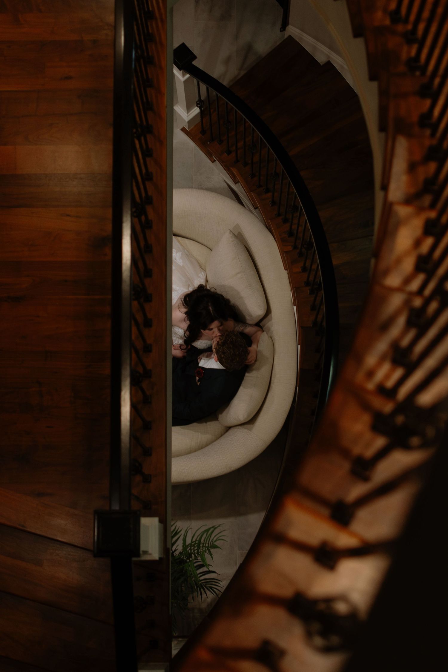 Aerial view of couple embracing on a round white sofa at the center of an elegant spiral staircase.