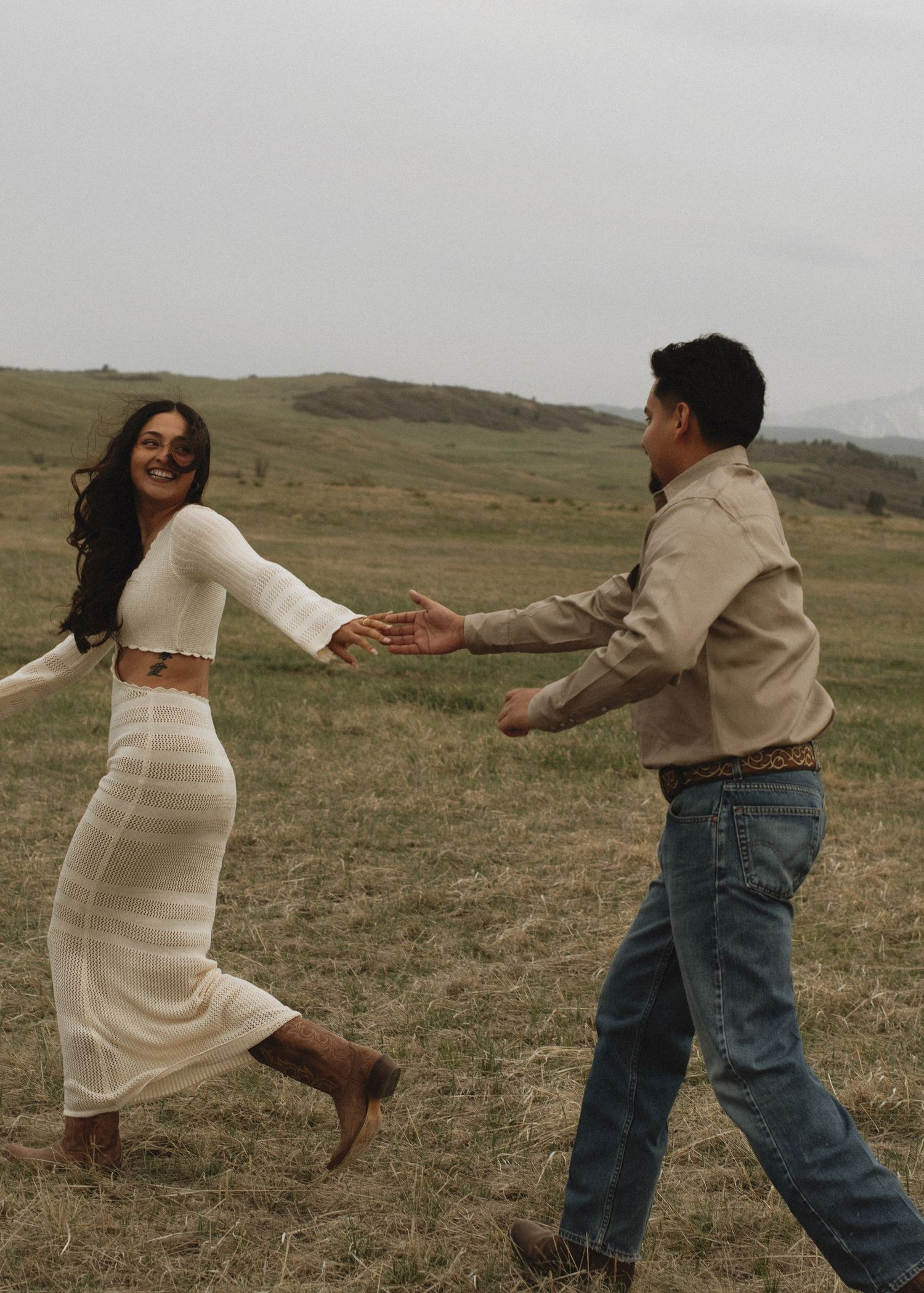 Joyful couple holds hands while running through open grassy field during outdoor engagement shoot.