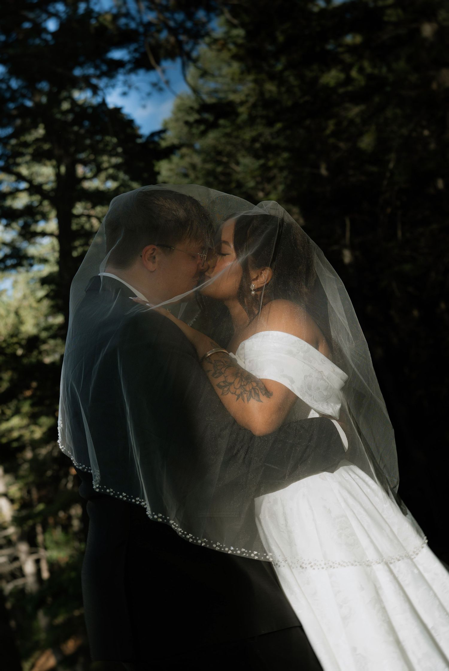 Bride and groom sharing a romantic kiss under a flowing white veil, surrounded by tall trees in an outdoor wedding setting.