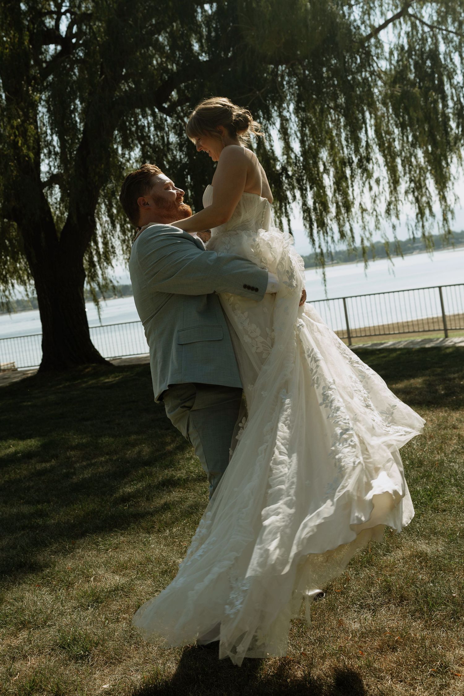 Groom lifting bride in flowing white gown under willow tree by lakeside on sunny wedding day.