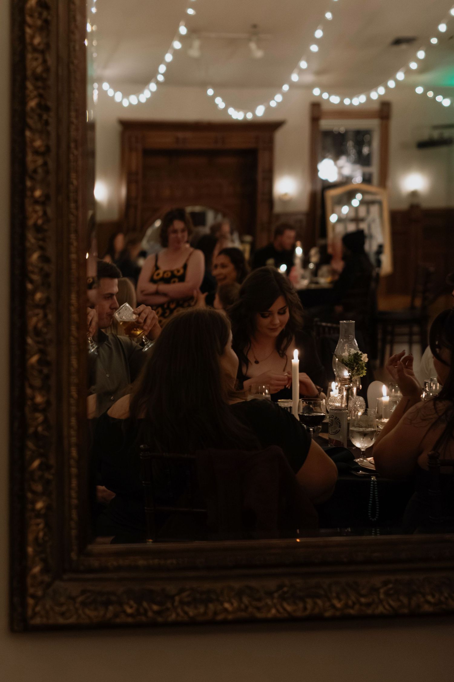 Guests dining by candlelight in an elegant restaurant reflected in an ornate gold mirror with string lights above.
